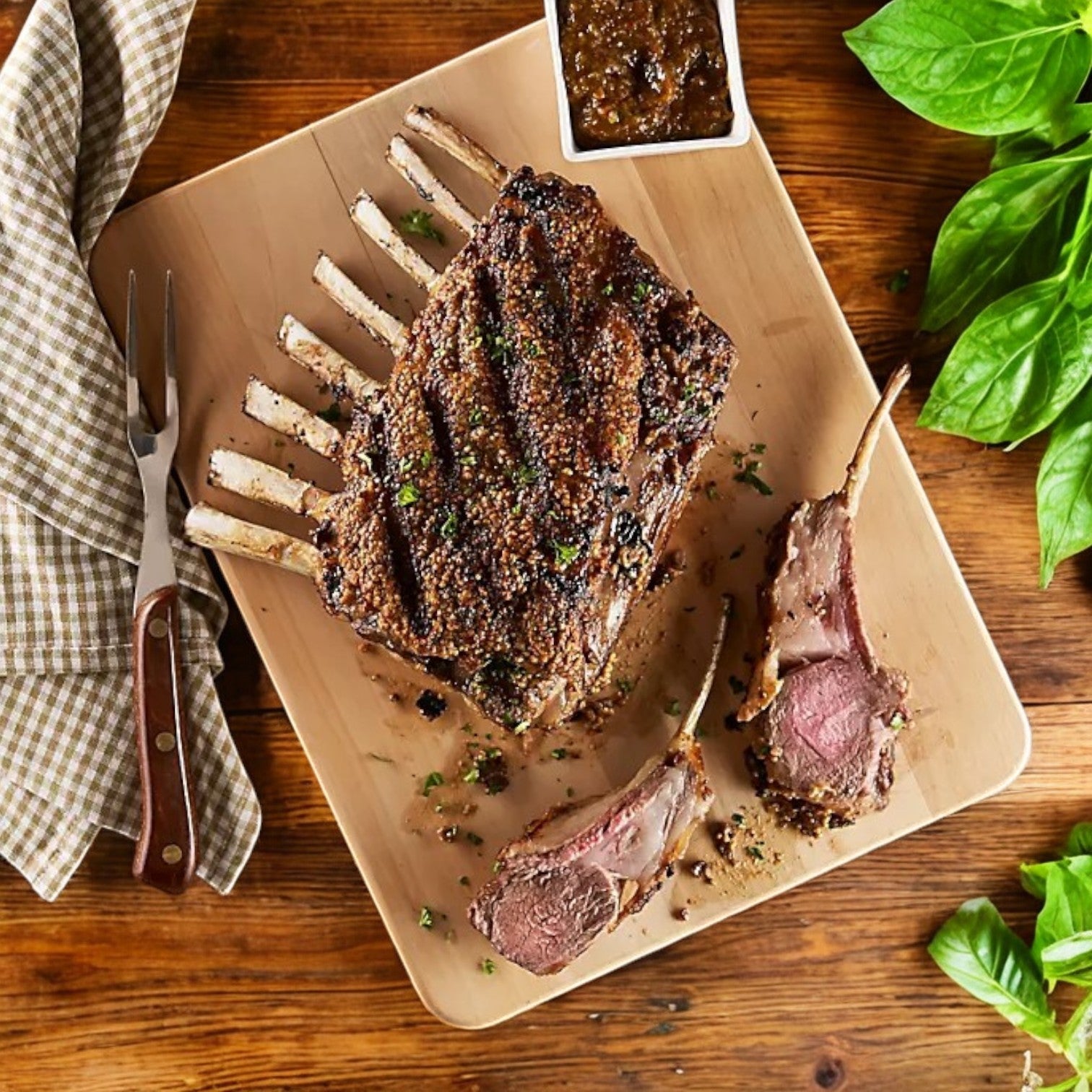 New Zealand Spring Lamb’s Halal Frenched Lamb Rack, 3 lb avg, 2 per pack, served on a wooden board with two succulent chops cut, a carving fork, basil garnish, checkered napkin, and a side of sauce.