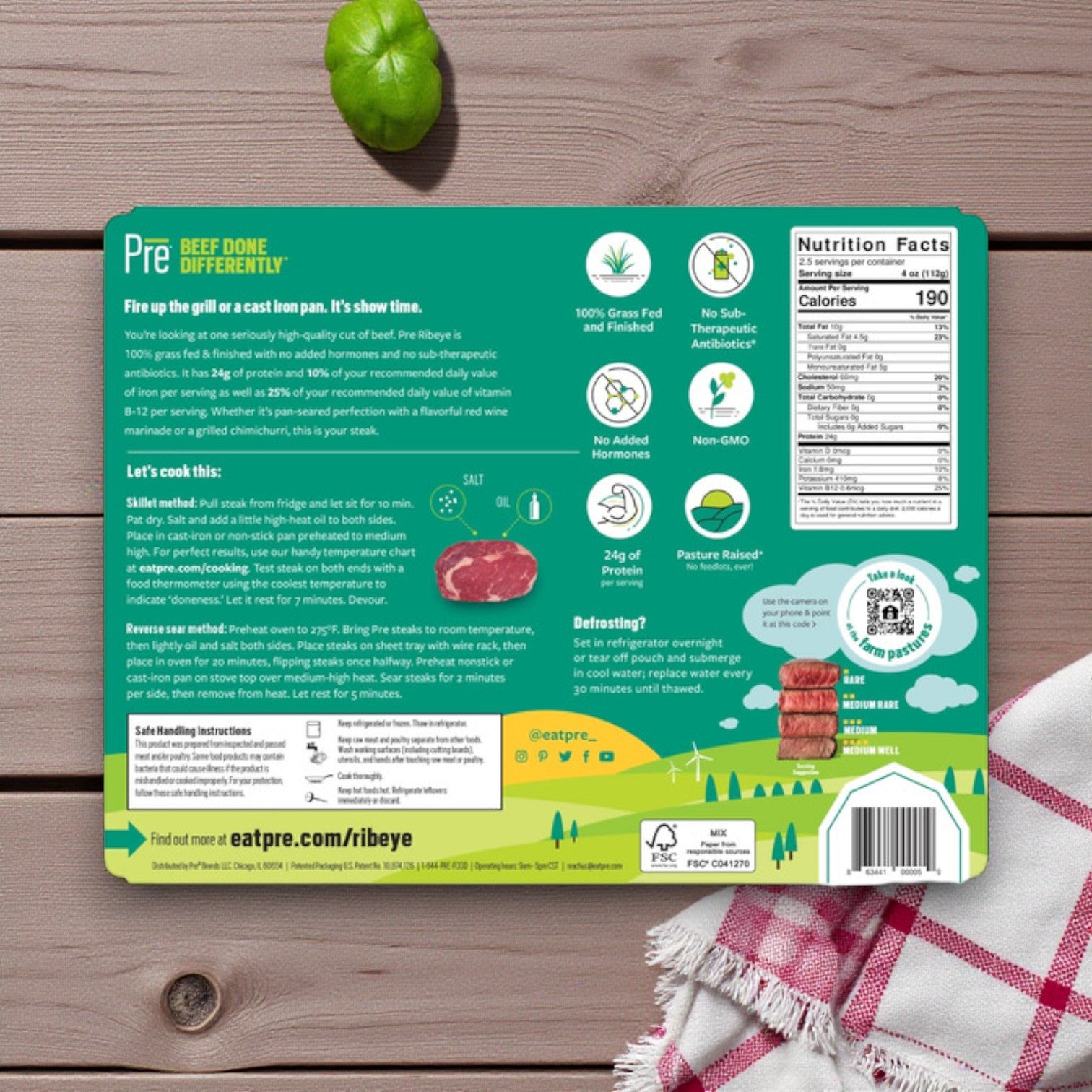 Back of Pre Brands Ribeye Steak- 100% Grass Fed Fresh Beef, 10 oz. packaging on a wooden surface with a green apple and red-and-white checkered cloth, showing cooking instructions, nutrition facts, and product details.