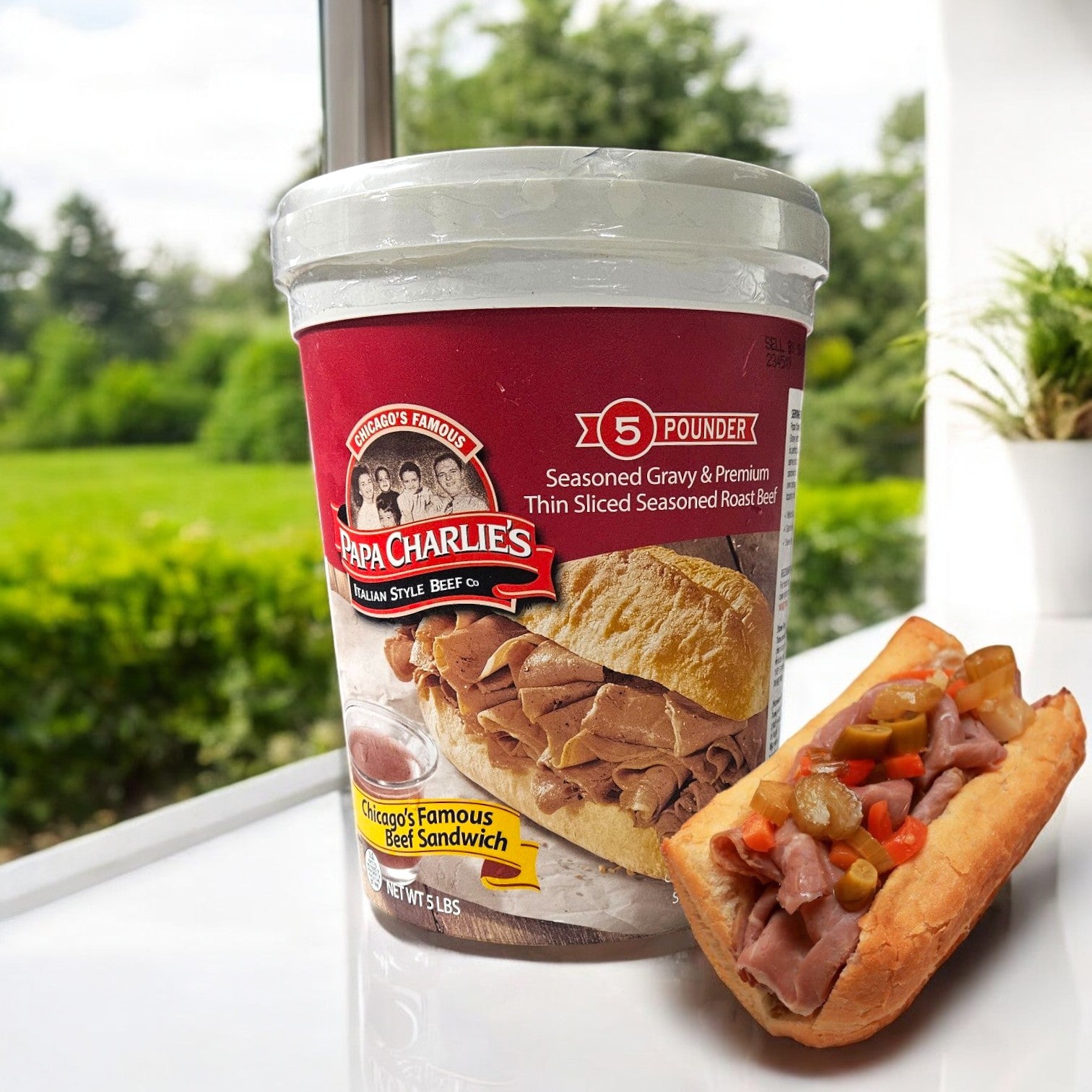 A package of Papa Charlie's Seasoned Gravy & Premium Thin Sliced Roast Beef rests beside a beef-and-pepper sandwich on a white surface, with an inviting outdoor blur in the background.