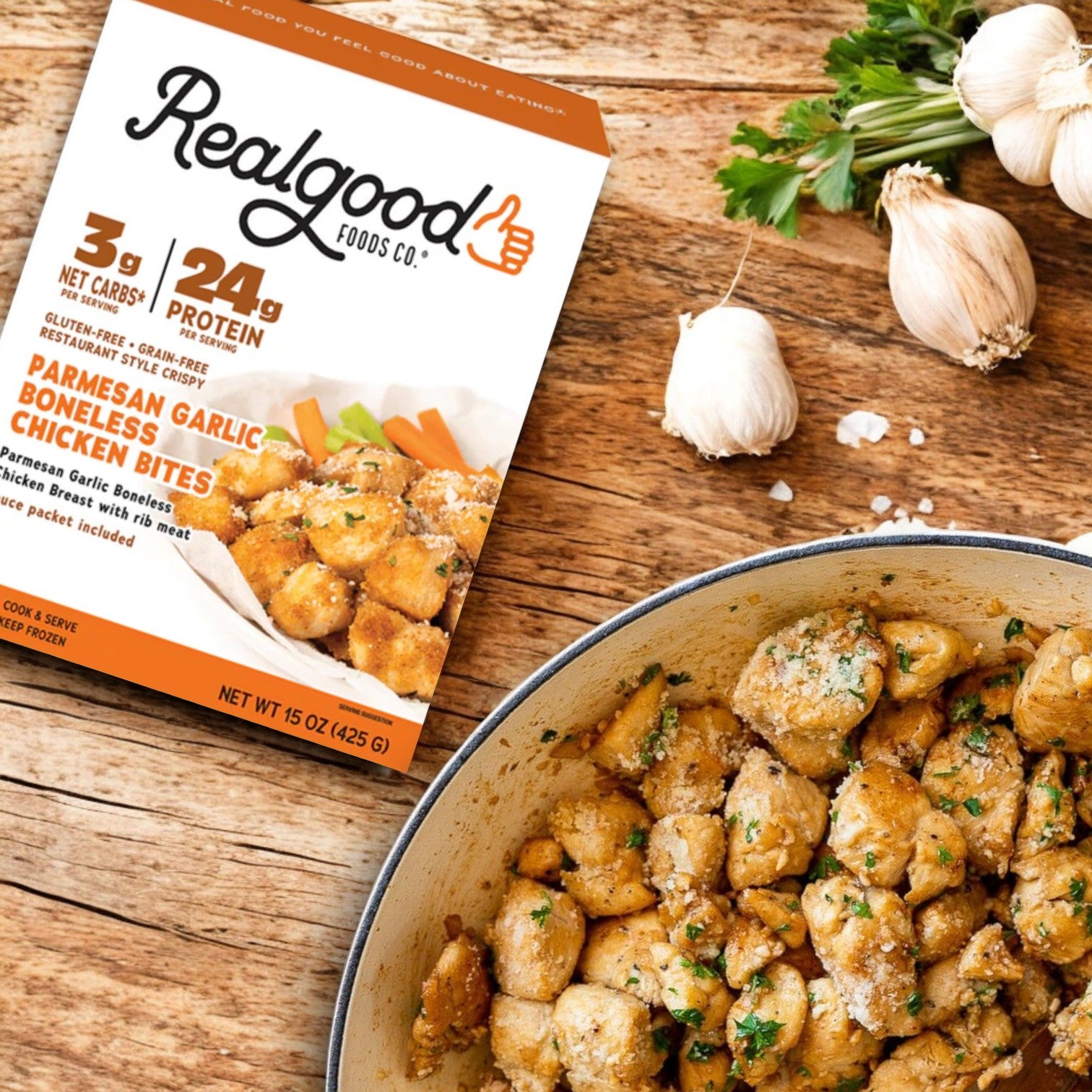 A box of Real Good Foods Low Carb Parmesan Garlic Boneless Chicken Bites (15 oz.), high-protein and gluten-free, sits beside a pan of herb-garnished cooked bites; garlic bulbs rest on the wooden table.
