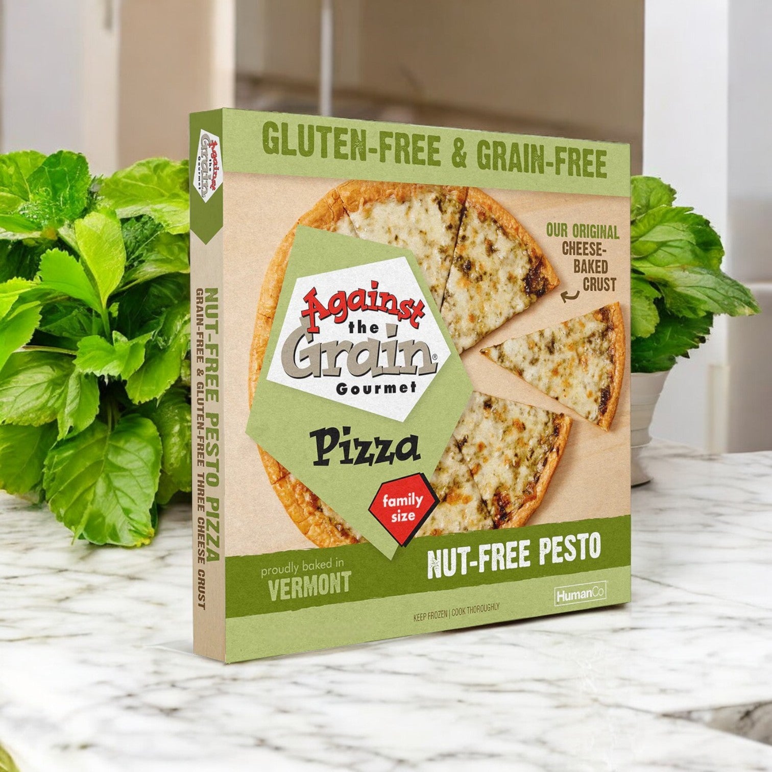 A 3-pack of Against the Grain Gourmet Pizza, Nut-Free Pesto, Family Size (22.4 oz), is shown on a kitchen counter with fresh green basil leaves in the background.