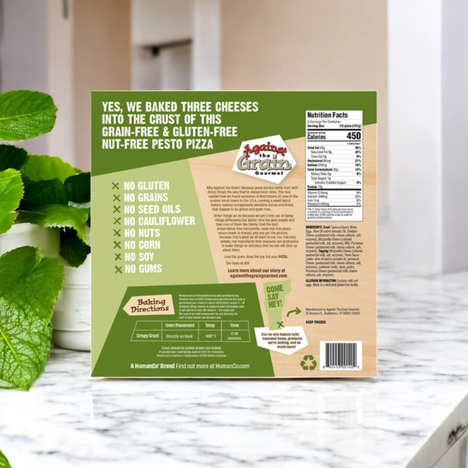 The back of an Against the Grain Gourmet Pizza, Nut-Free Pesto, Family Size box shows ingredients, nutrition facts, and features on a kitchen counter with green plants in the background. This is a gluten-free pizza in a 22.4 oz 3-pack.