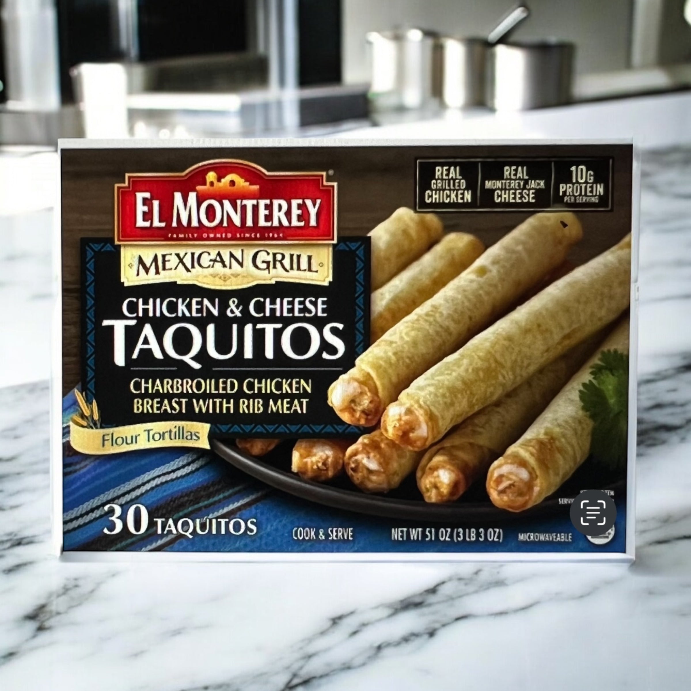 A box of El Monterey Mexican Grill Taquitos, Chicken & Cheese – 30 Count (51 oz), sits on a marble counter, featuring appetizing taquito images and product details on the colorful El Monterey packaging.