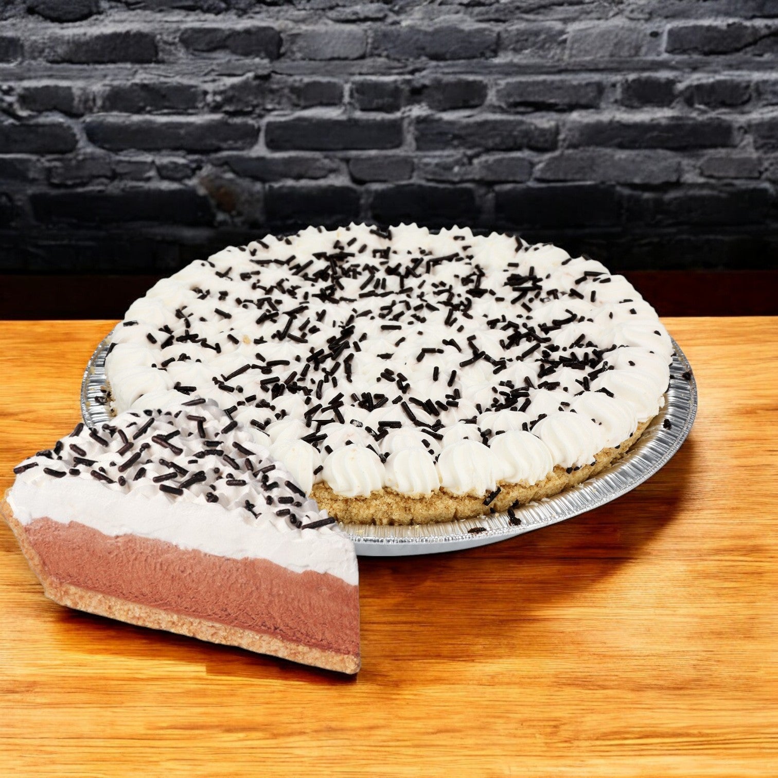 A Sysco Classic Chocolate Cream Pie with rich filling, whipped cream, and sprinkles sits on wood with a slice cut out, ready to thaw and serve. The 10" pie is set against a gray brick wall background.
