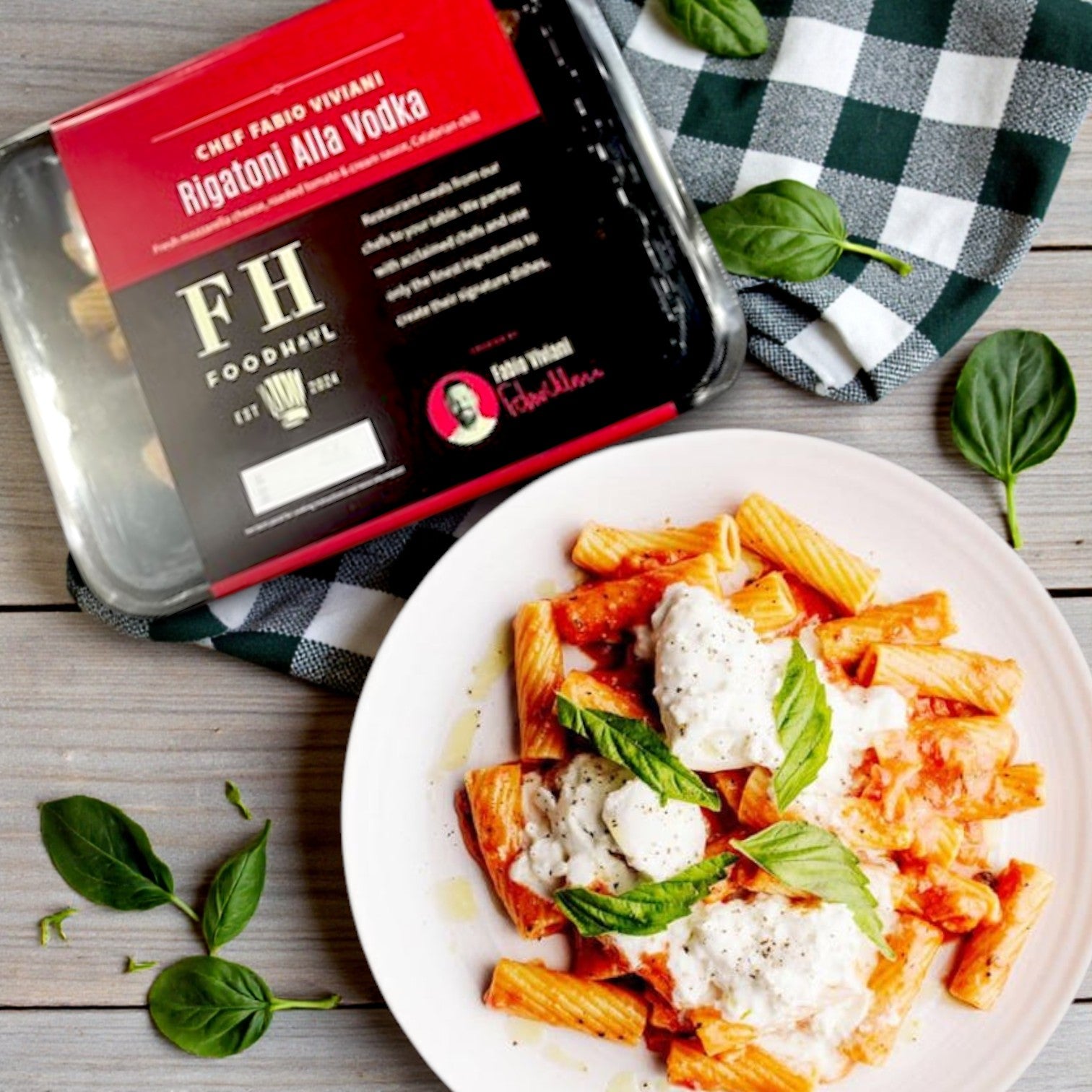 A box of FoodHaul Frozen Meals—Signature Chef Meal Combo with 5 Gourmet Entrées sits beside a plated serving, topped with basil and cheese, on a wooden table with spinach leaves and a checkered napkin.