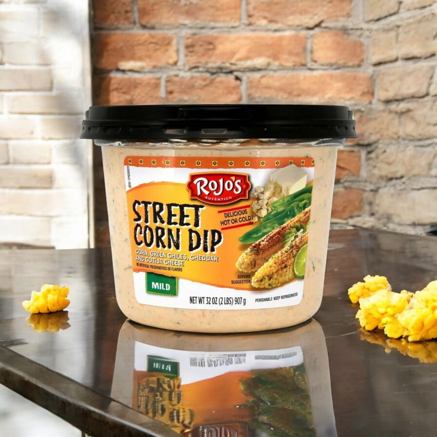 A 2-lb container of Rojo's Street Corn Dip sits on a reflective table with scattered corn kernels, highlighting its bold Mexican flavor; a brick wall is in the background.