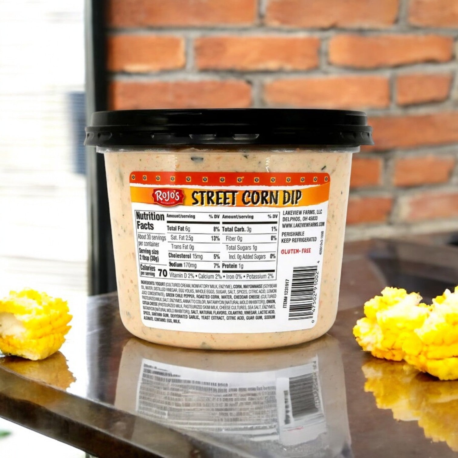 Rojo’s Street Corn Dip (2 lbs) by Rojo’s is shown with its nutrition label on a metal surface, corn on each side, and a brick wall background—perfect as a flavorful party snack inspired by bold Mexican street corn.