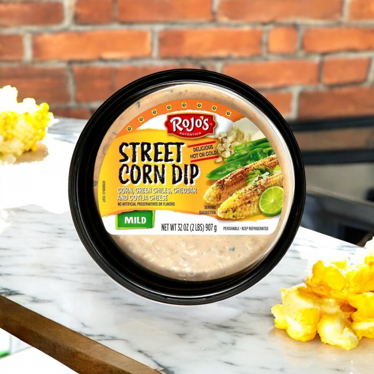 A 2-lb container of Rojo’s Street Corn Dip by Rojo’s, labeled “Mild,” sits on a marble surface with corn and chips nearby—offering vibrant Mexican flavor for your next party—while a brick wall adds rustic charm in the background.