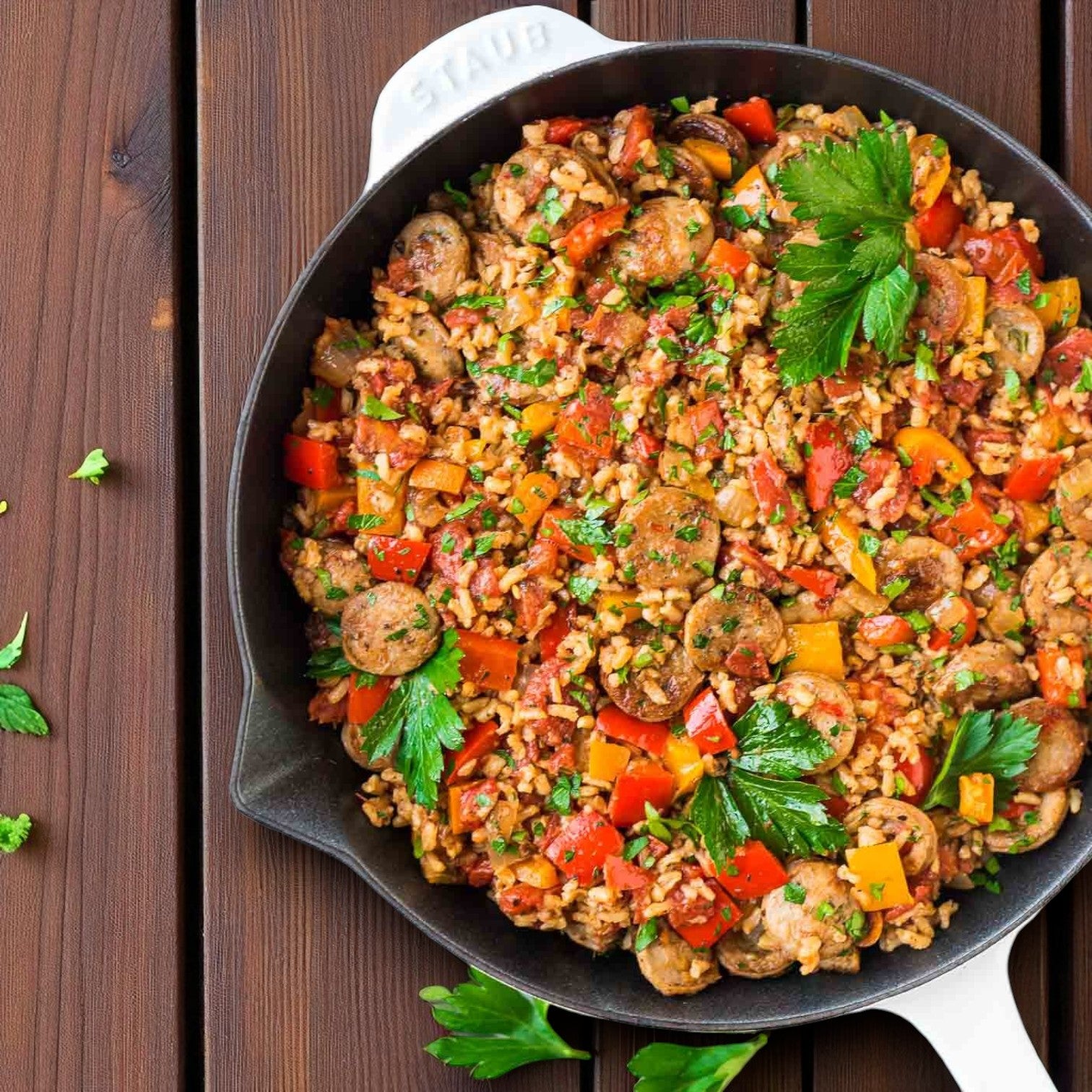 A skillet of rice, Sysco’s Fontanini Mild Italian Pork Sausage (Raw Bulk 20 lbs), sliced and cooked with red and yellow bell peppers, tomatoes, and parsley, is served on a wooden table.