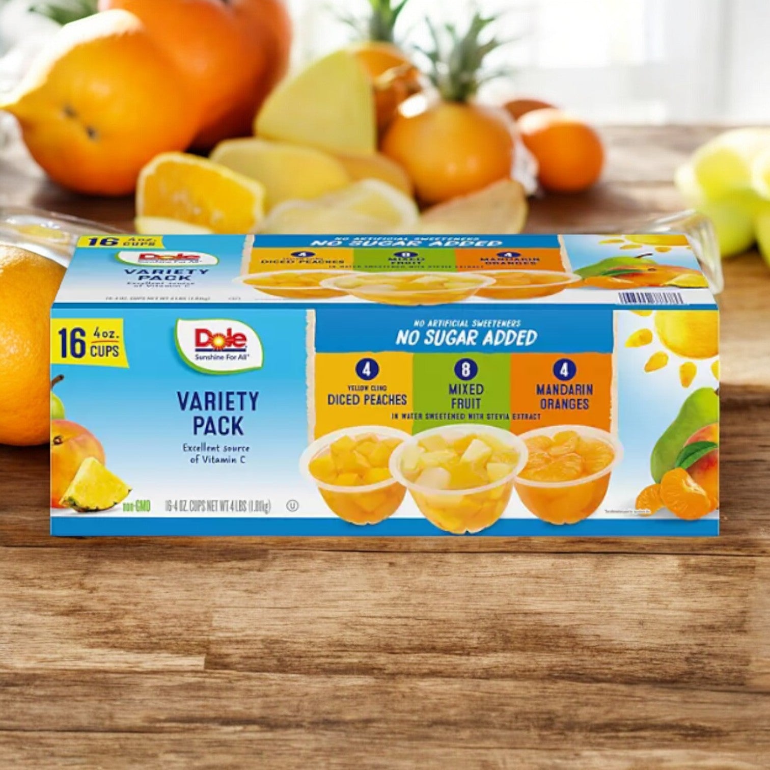 A 16-pack of Dole No Sugar Added Mixed Fruit Variety Pack 4 oz cups sits on a wooden table with fresh citrus fruits in the background. The packaging highlights gluten-free snacks, no sugar added, and three delicious fruit cup flavors.