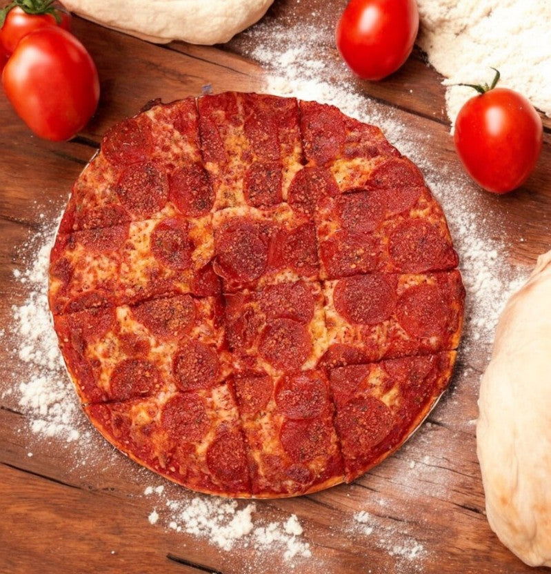 A Labriola Chicago Pepperoni Thin Crust Pizza-1 Count from Labriola Chicago rests on a wooden surface, topped with savory ingredients, and surrounded by fresh tomatoes, dough, and flour.