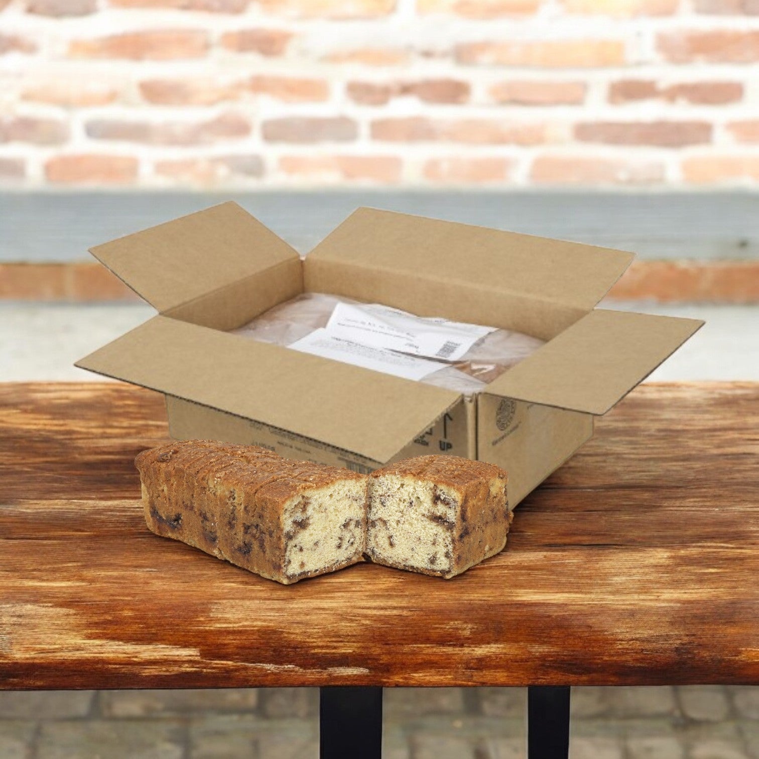 On a wooden table by a brick wall, there is an open cardboard box filled with papers next to a Sysco Cinnamon Streusel Sliced Cake Loaf (2 loaves of 12 inches, totaling 36 pieces).