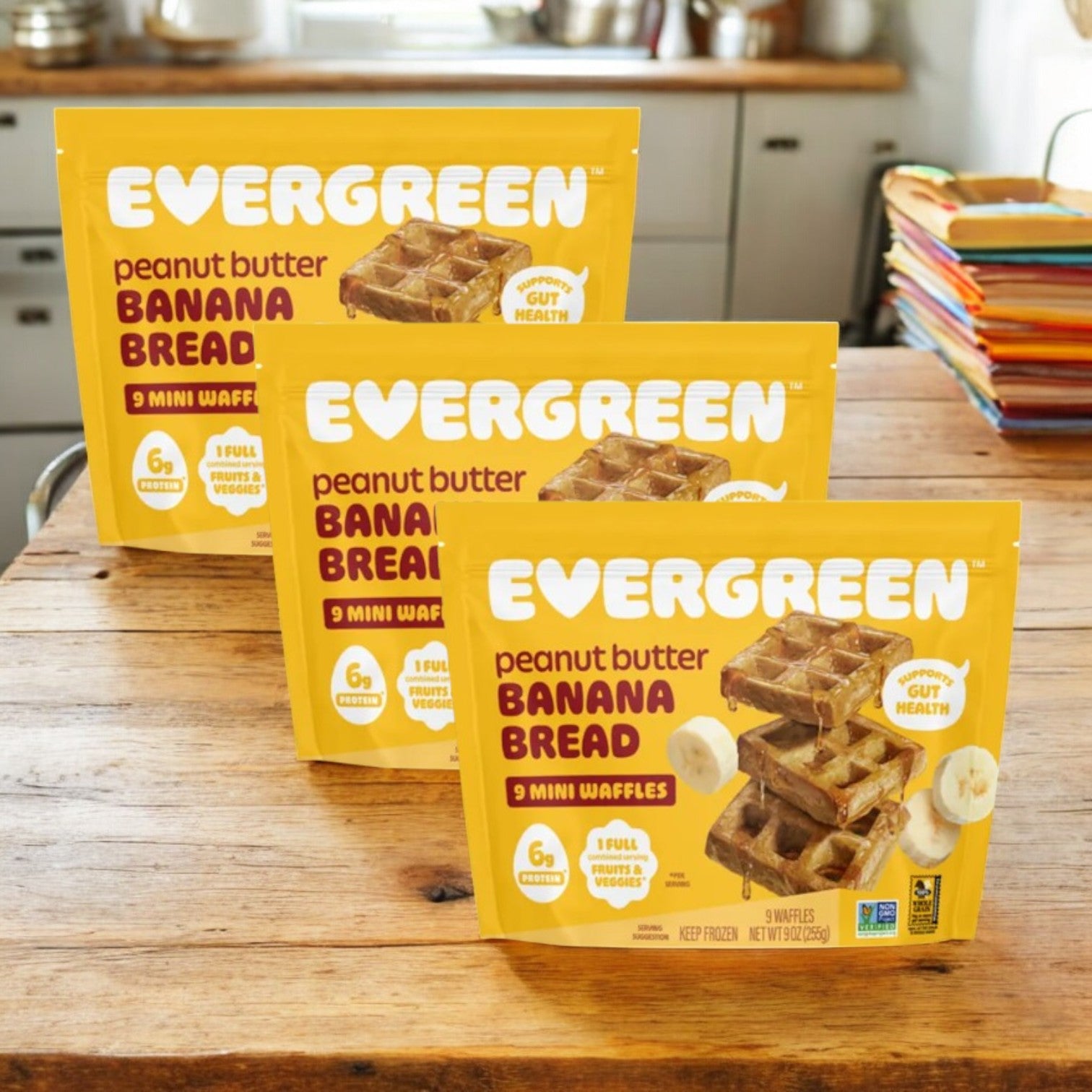 Three packs of Evergreen Peanut Butter Banana Bread Frozen Mini Waffles, 9 oz. each, are displayed on a wooden table in a cozy kitchen, ready to be enjoyed as a tasty breakfast or snack.