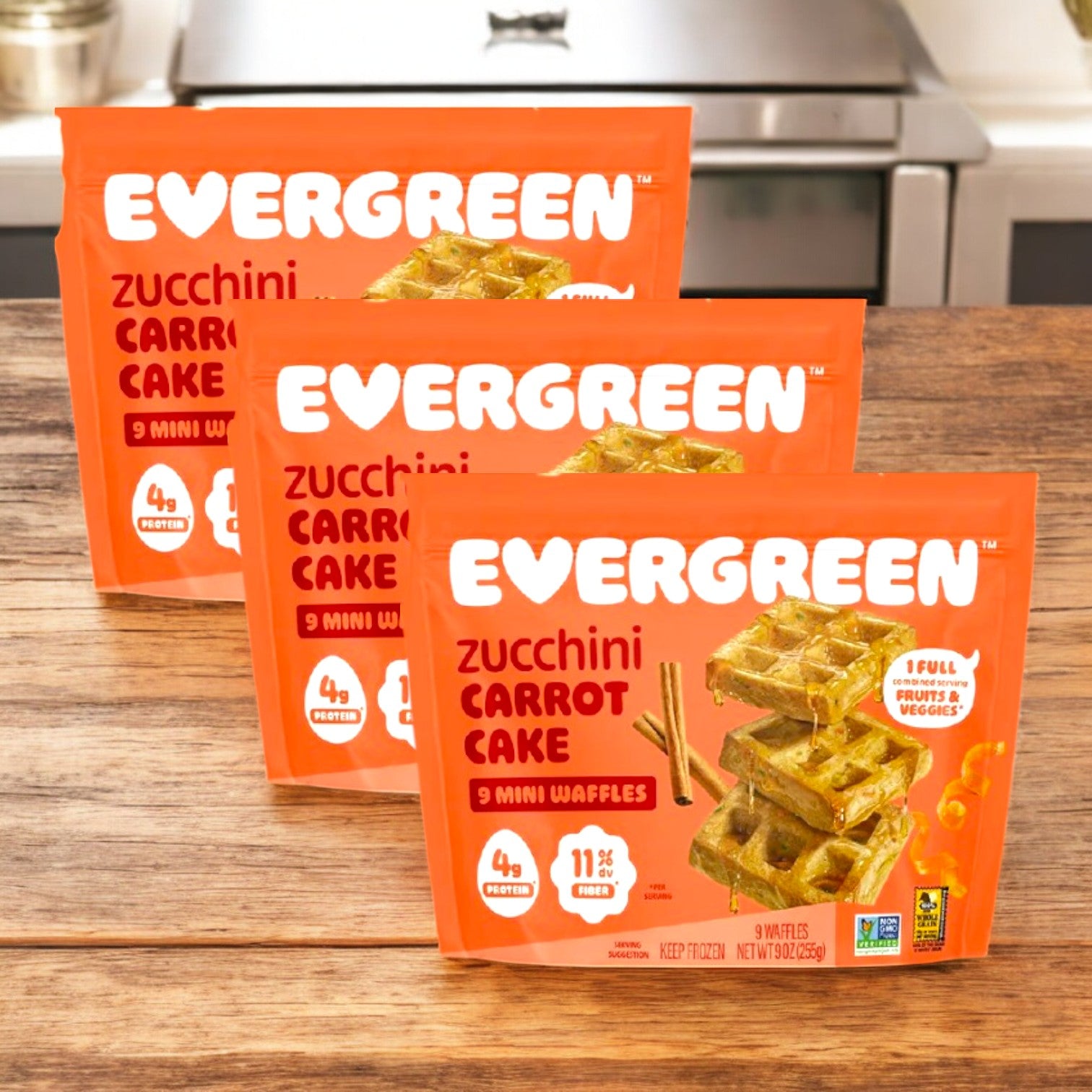 Three packs of Evergreen Zucchini Carrot Cake Frozen Mini Waffles (9 oz., 3 Count) are displayed on a wooden surface, highlighting these tasty and nutritious breakfast treats from Evergreen.