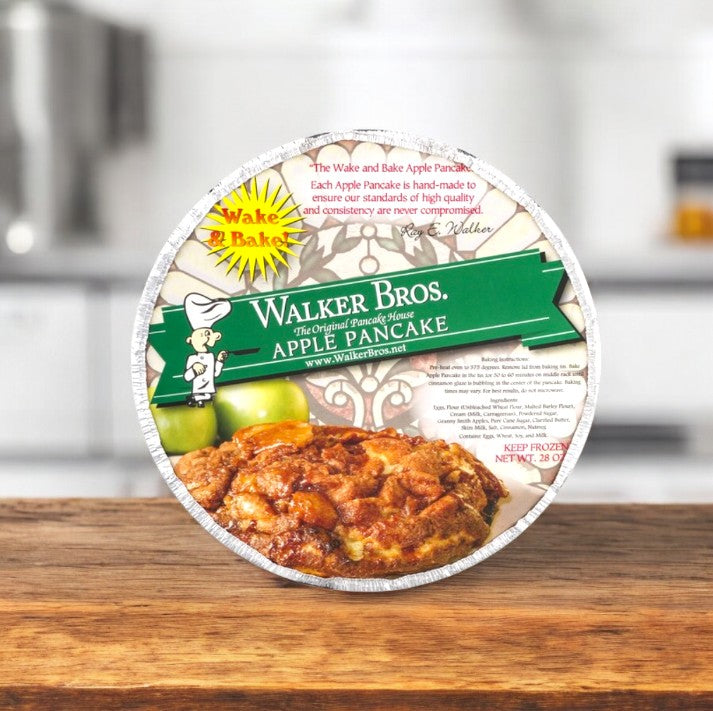A 28 oz Walker Bros, The Original Pancake House Apple Pancake sits in a round foil container with green Walker Bros branding and a pancake photo, displayed on a wooden surface in a cozy kitchen.
