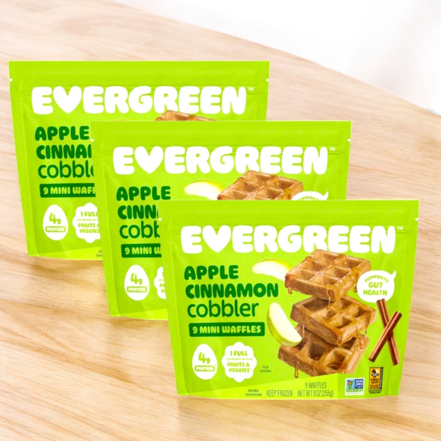 Three Evergreen Apple Cinnamon Cobbler Frozen Mini Waffles 9 oz packs are displayed on a wooden surface, showing the brand, product details, and images of mini waffles on the packaging front.