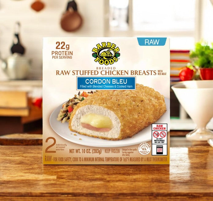 Barber Foods Breaded Raw Cordon Bleu Stuffed Chicken Breasts, 2 Count pack, on a wooden table filled with ham and Swiss cheese, set against a blurred kitchen background.
