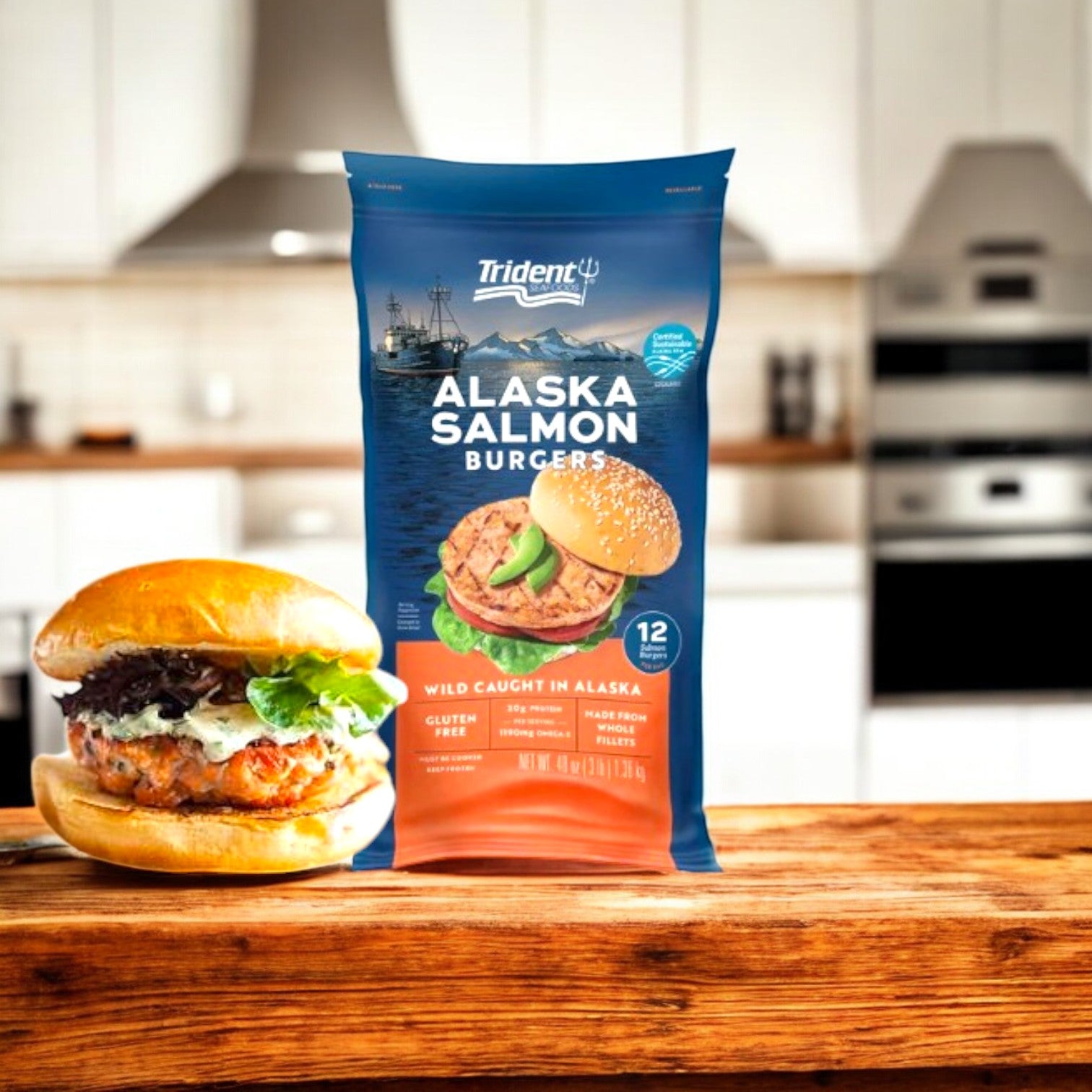 A 48 oz box of Trident Seafoods Alaskan Salmon Burgers stands on a wooden surface beside a cooked salmon burger; both are rich in Omega-3s, with a modern kitchen in the background.