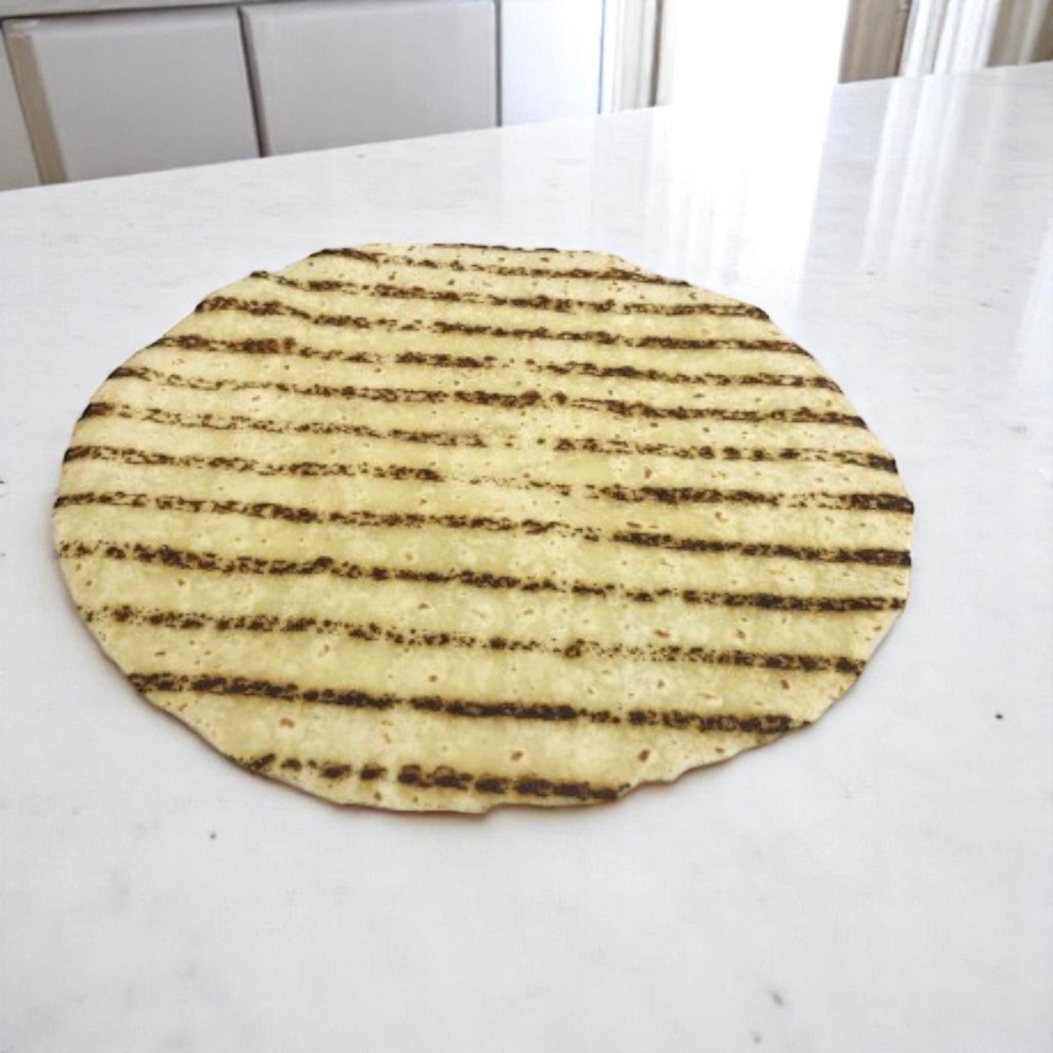 A single Easy Lunches Kontos Tortilla Wrap Gluten Free Rice Flour 12 Inch Grilled sits on a white marble countertop, showcasing these tasty wheat-free wraps—available in 6 packs, totaling 72 wraps—as a delicious gluten-free alternative.