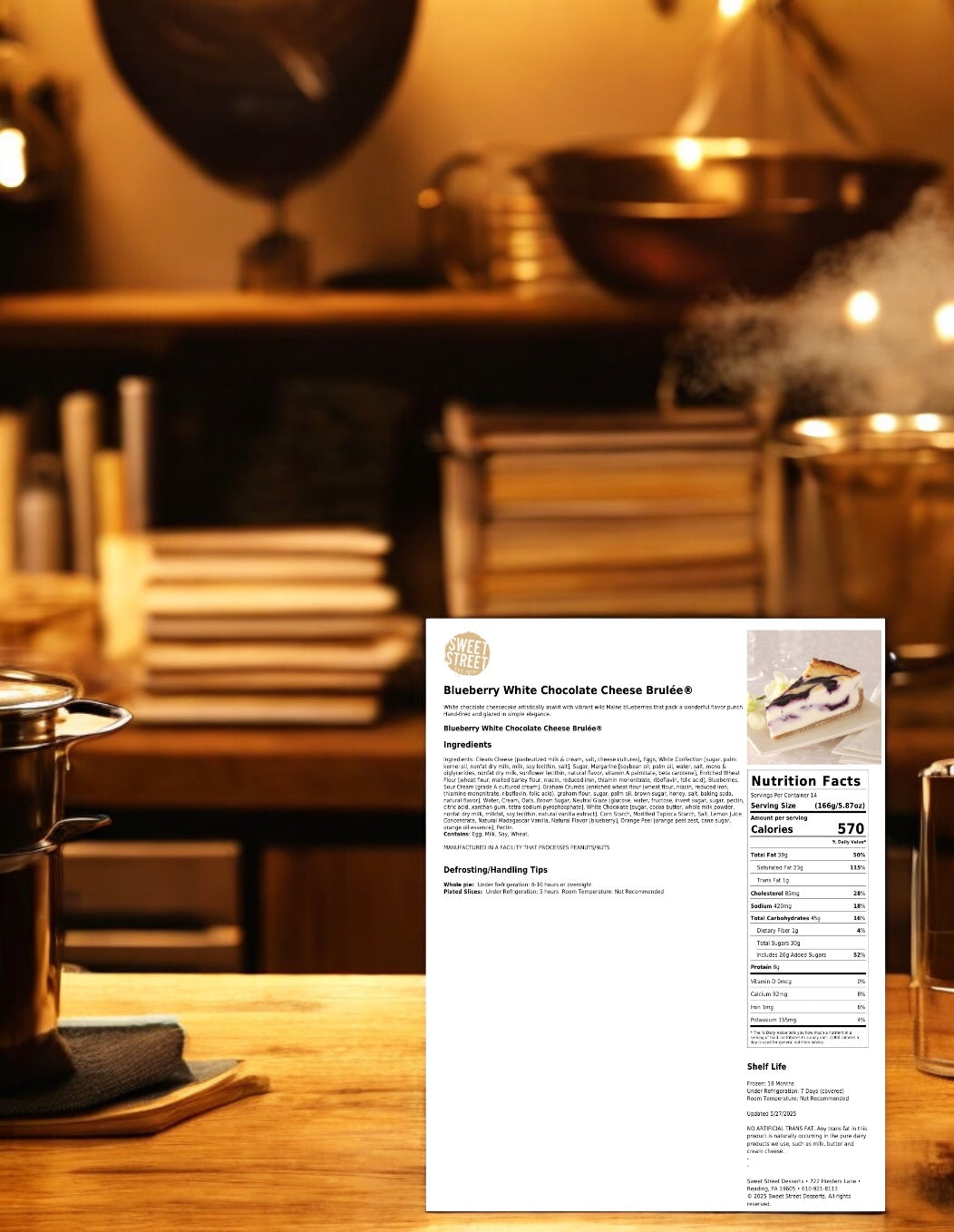 A printed recipe for Sweet Street Blueberry White Chocolate Cheese Brulée 2/83 OZ is in focus against a blurred kitchen scene with pots and stacked plates, featuring the gourmet dessert and brand Sweet Street, ready to serve.