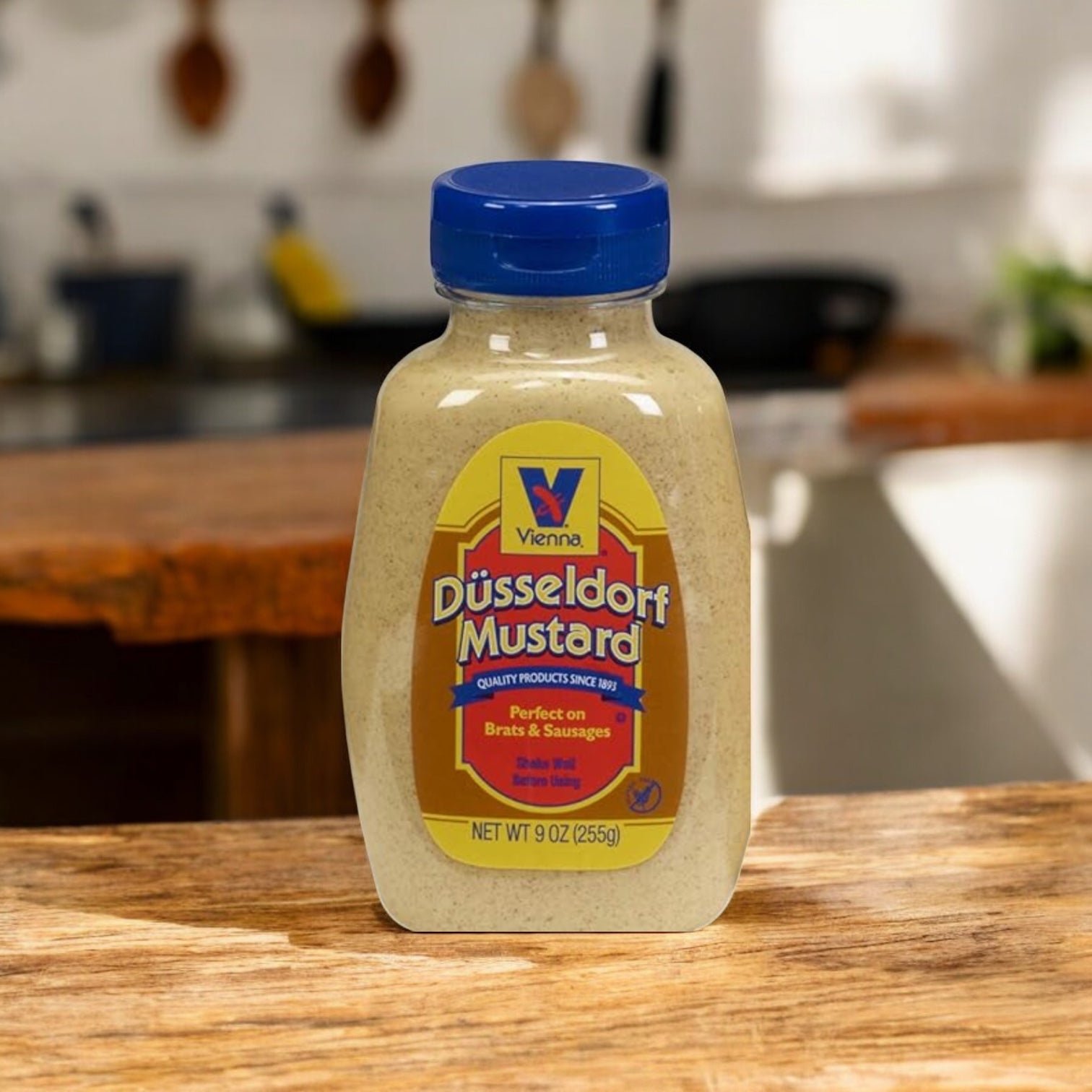 A 1-gallon jar of Easy Lunches Vienna Dusseldorf Mustard is displayed on a wooden surface in a kitchen, with natural light streaming in from the background.