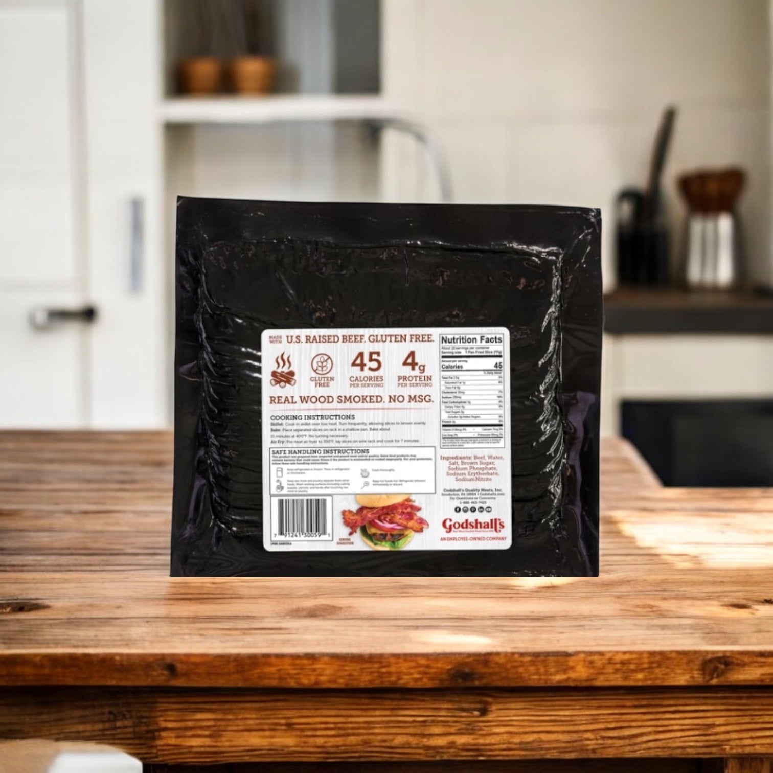 A vacuum-sealed package of Easy Lunches Godshall's Beef Bacon 20 oz., 3 count, rests on a wooden countertop in a kitchen with white cabinets and assorted kitchen items in the background.