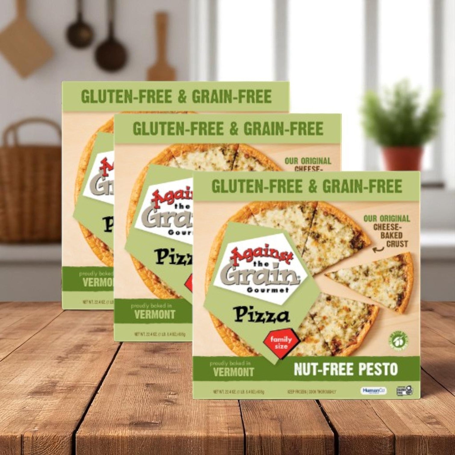 Three packs of Against the Grain Gourmet Pizza, Nut-Free Pesto, Family Size (22.4 oz each), are displayed on a wooden kitchen table—ideal for an allergen-friendly meal.