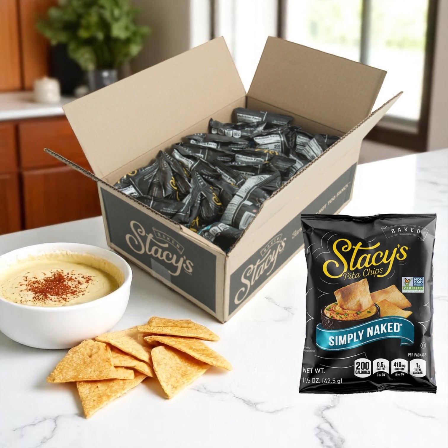 A box of Easy Lunches Stacys Pita Chips Large Single Serving Simply Naked 24 Case sits on the kitchen counter beside a bowl of hummus, pita chips on a plate, and an individually packaged snack-size bag.
