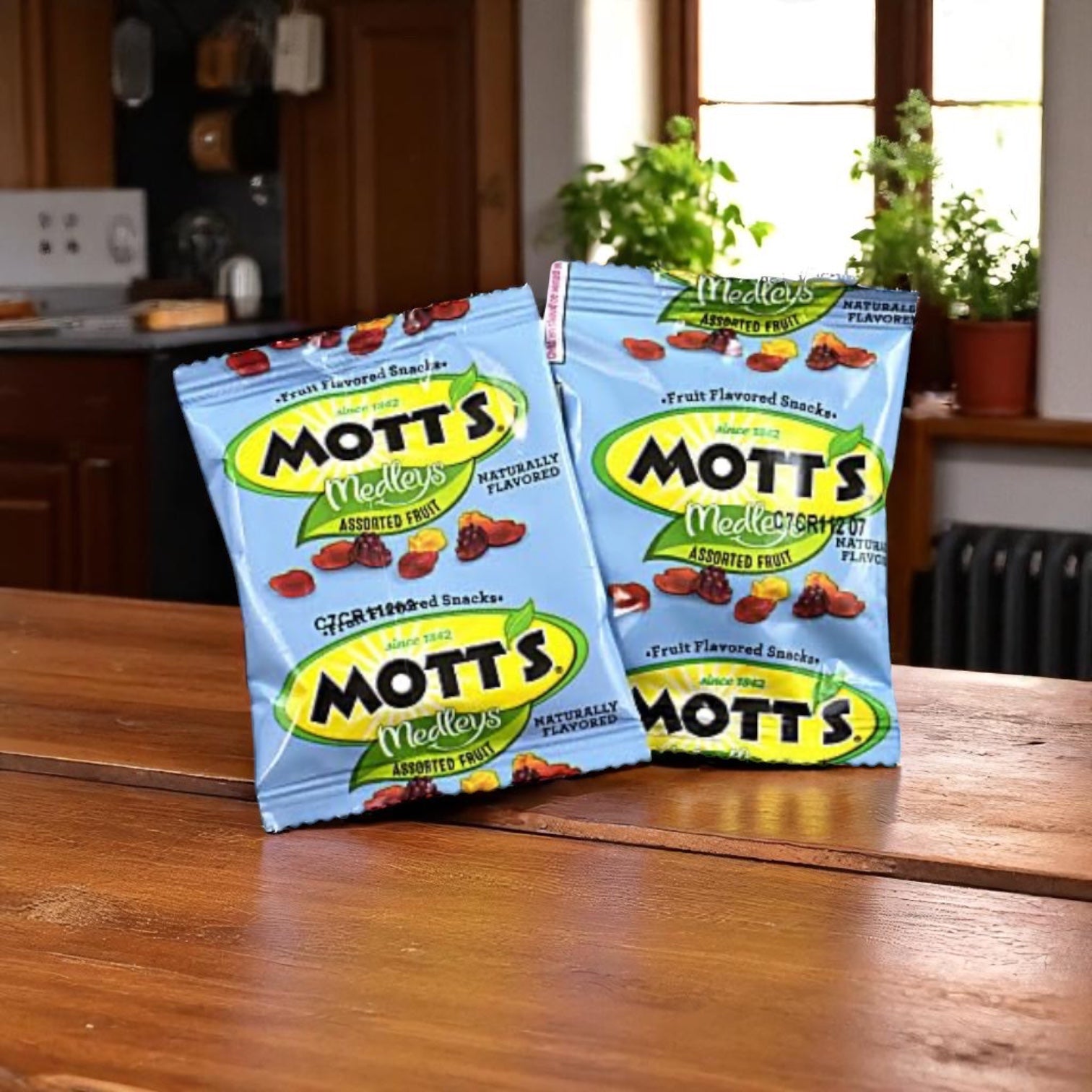 Two packs of Easy Lunches Mott's Snack Fruit Gummies Assorted Fruit, 1.6 oz, sit on a wooden kitchen table with plants by the window in the background.
