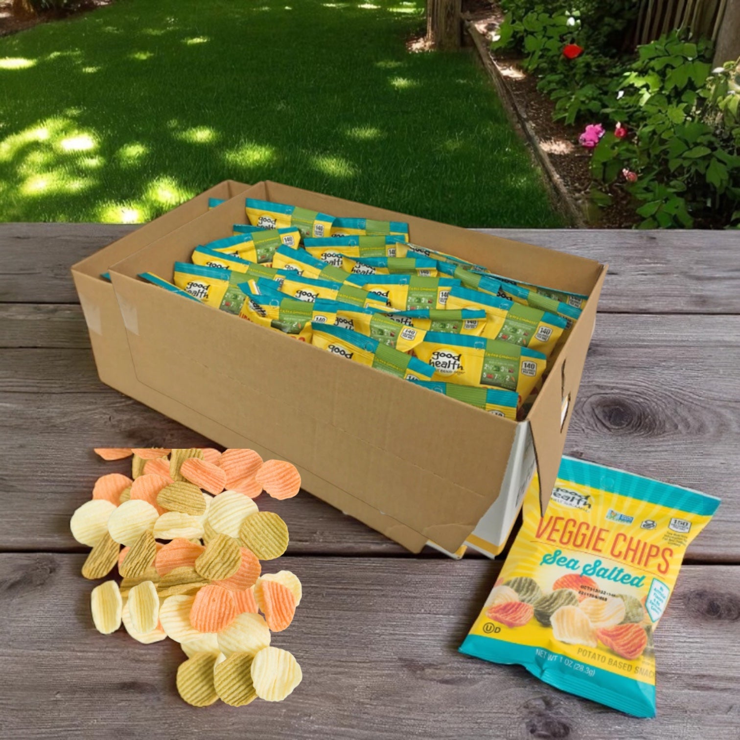 A cardboard box of Easy Lunches Good Health Vegetable Chip Snack, 1 oz. - 24 a case, sits on a wooden table outdoors. An open bag and some veggie chips are nearby, with a grassy yard in the background.