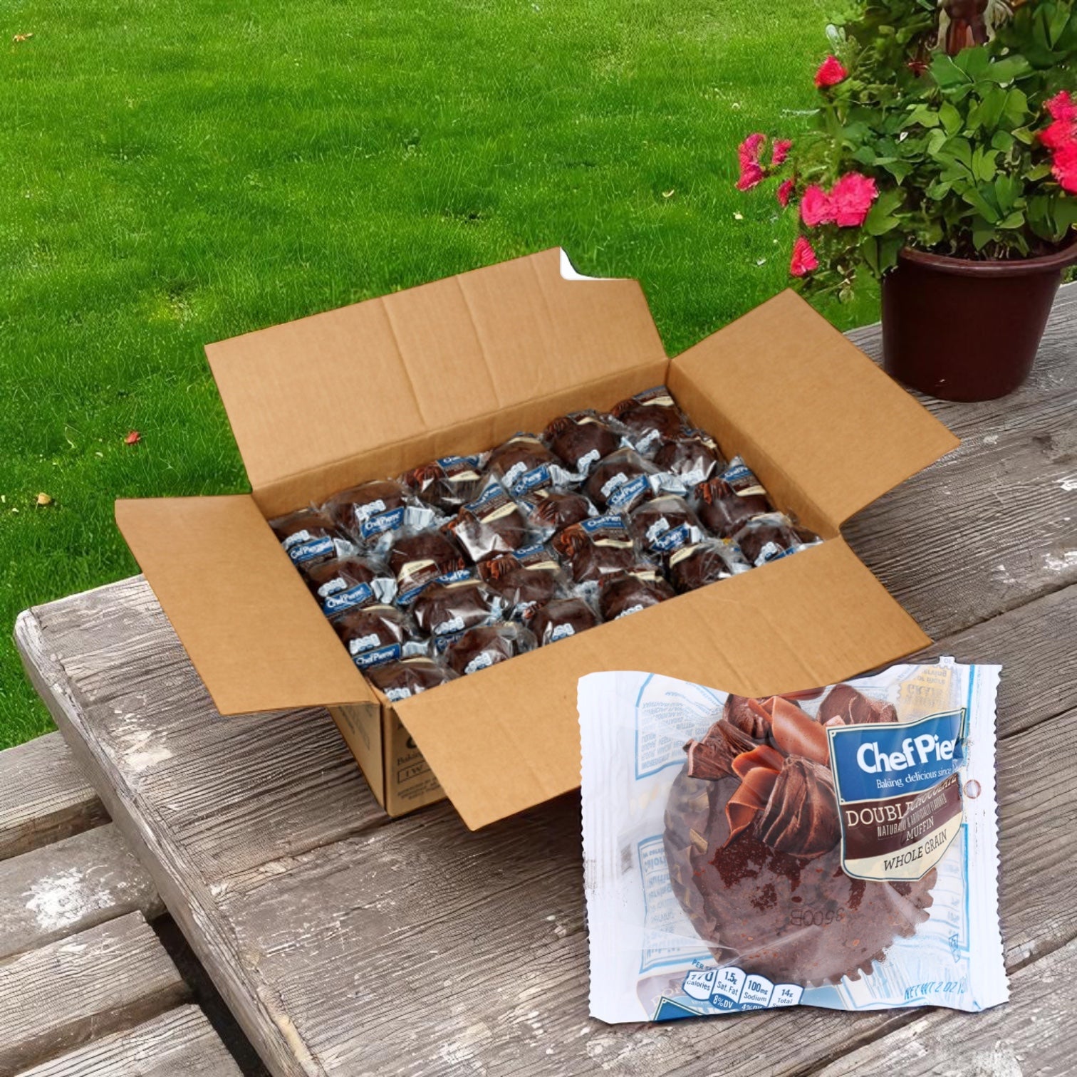 An open box of Easy Lunches Chef Pierre Muffin Chocolate Double Whole Grain 2 oz 48 Pack sits on a wooden picnic table outdoors next to a potted plant with pink flowers, each muffin individually wrapped and filled with semi-sweet chocolate chips.