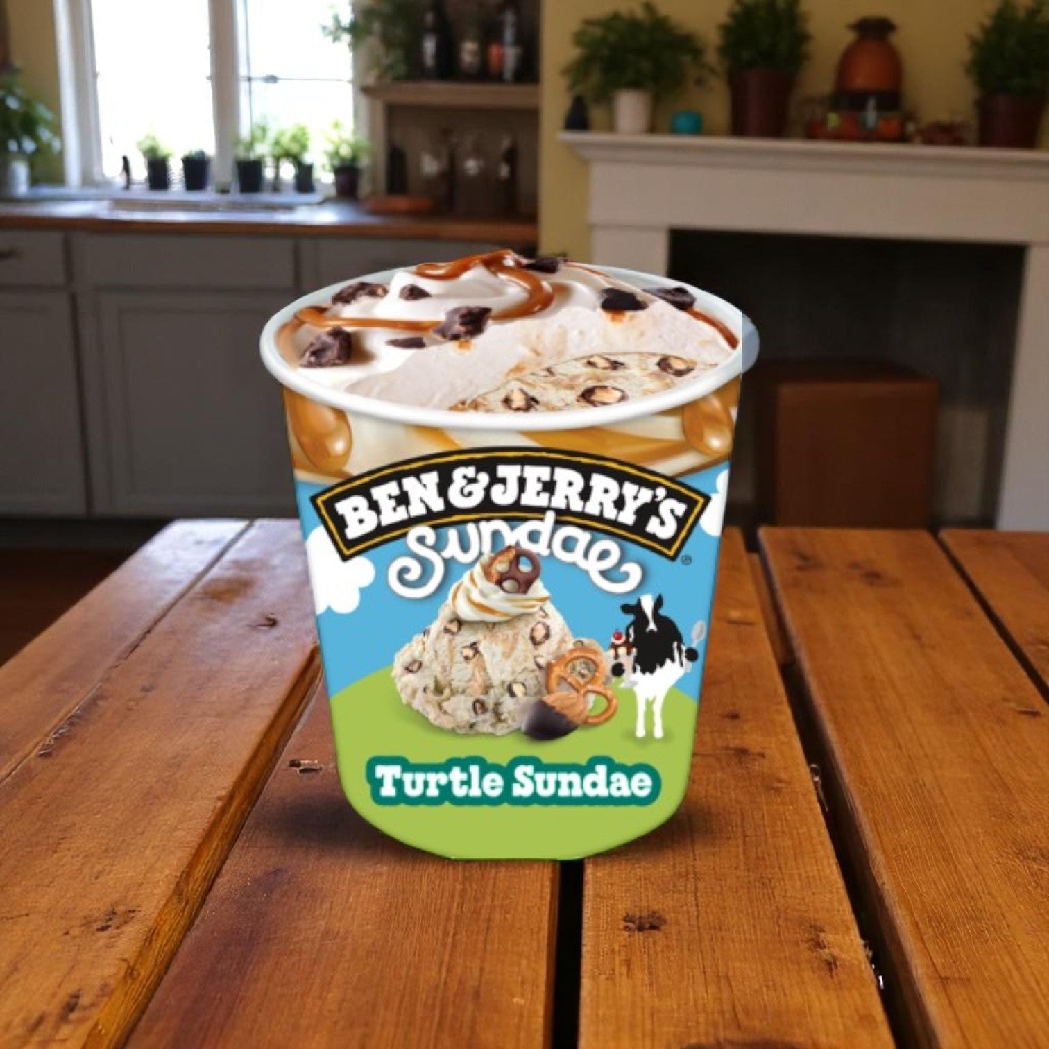 A pint of Ben & Jerry's Turtle Sundae Buttery Ice Cream (14.4 FL Oz) rests on a kitchen table, surrounded by plants and a fireplace, highlighting the indulgent flavor from Ben & Jerry’s.