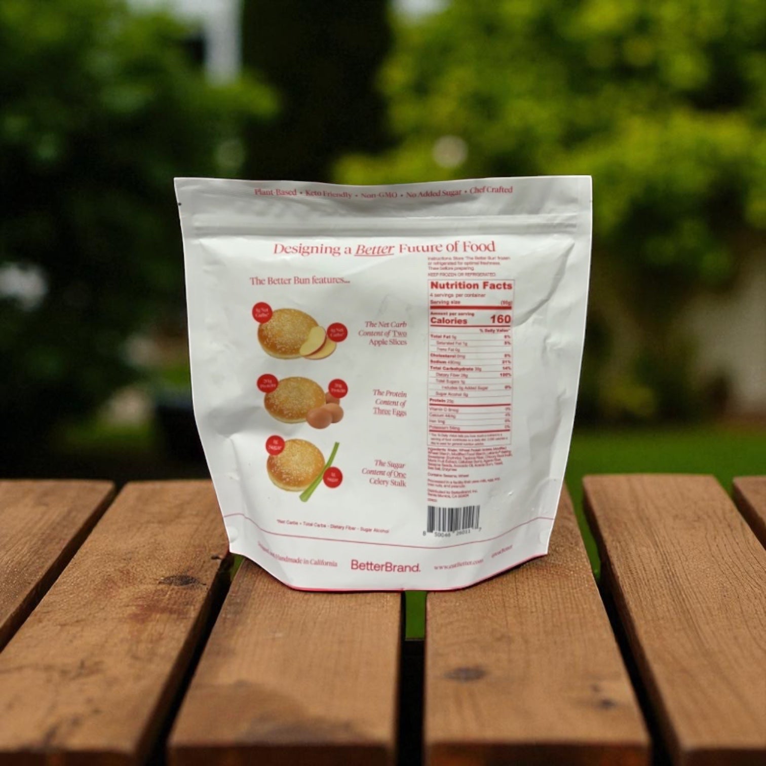 A bag of BetterBrand The Better Bun – Sesame Style (1 bag, 4 buns) sits on a wooden table outdoors, displaying its back label with nutrition facts. Green foliage appears blurred in the background. In stock and ready to ship!.
