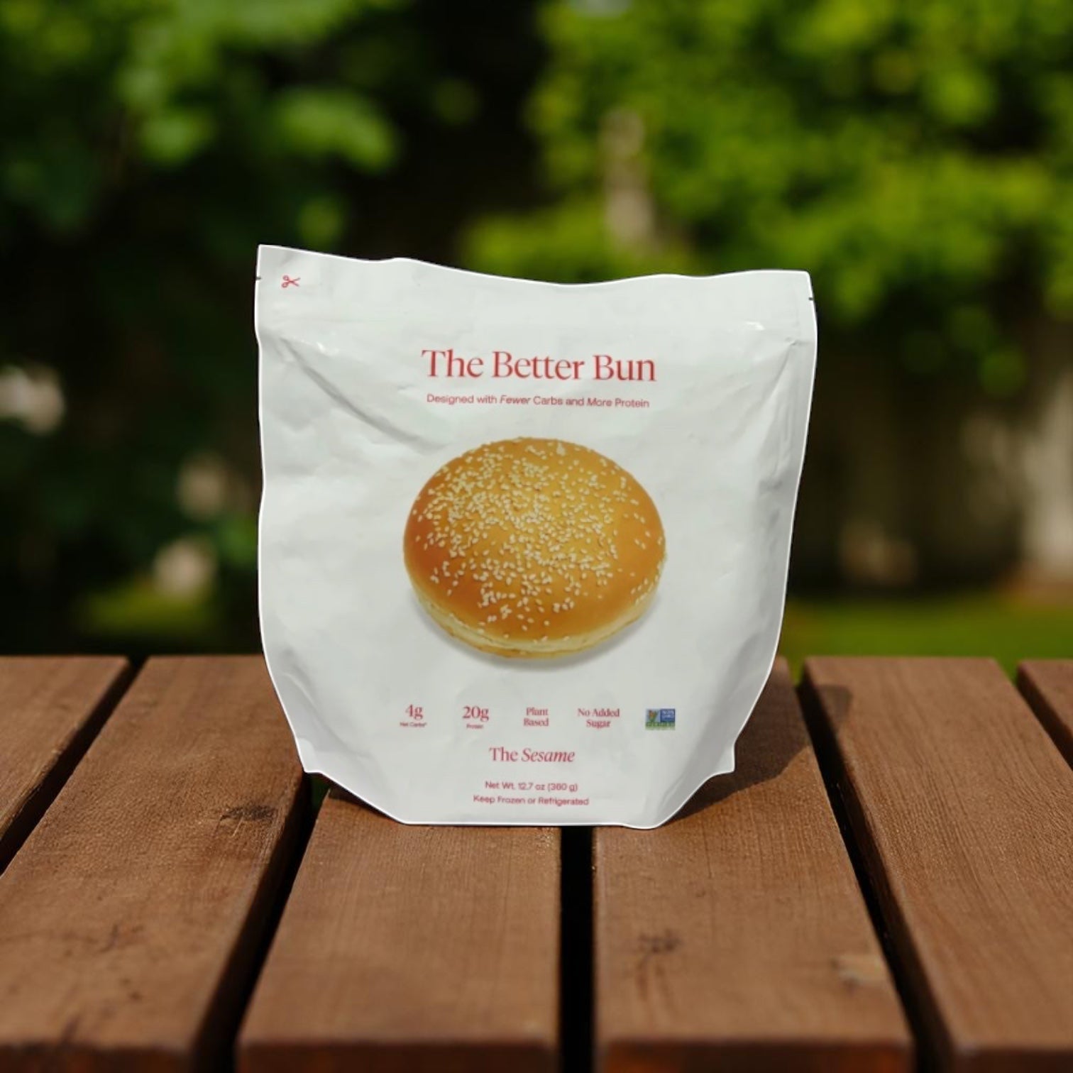 A white package labeled "BetterBrand, The Better Bun – Sesame Style" (1 Bag, 4 Buns) sits on a wooden surface with softly blurred greenery in the background. In stock and ready to ship!.