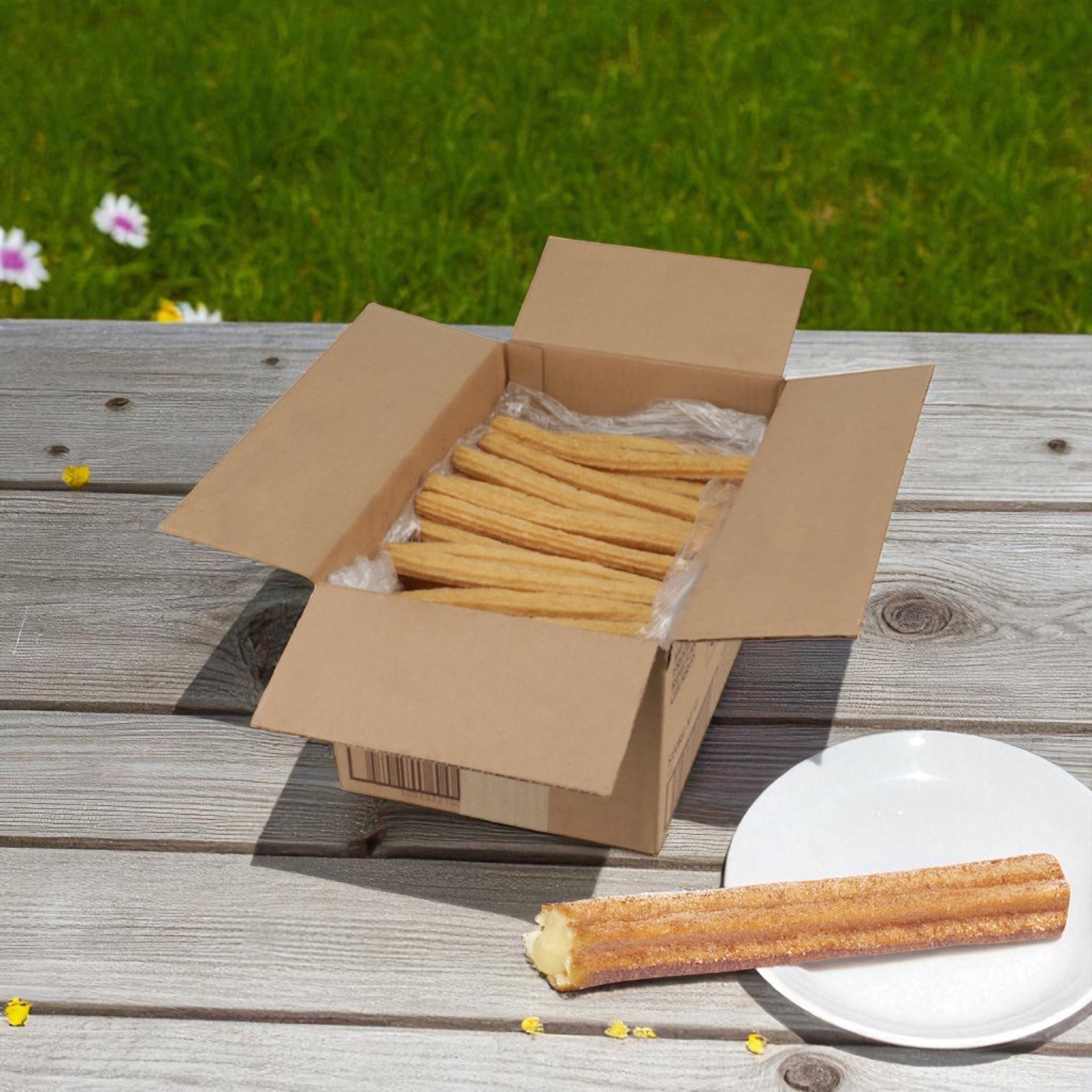 A box of Hola Churro® – Churro Prefried Bavarian Cream (100 count 10 pack) by Hola Churro sits on a wooden table outdoors, with a plate featuring one churro beside it, framed by grass and flowers for an inviting scene.