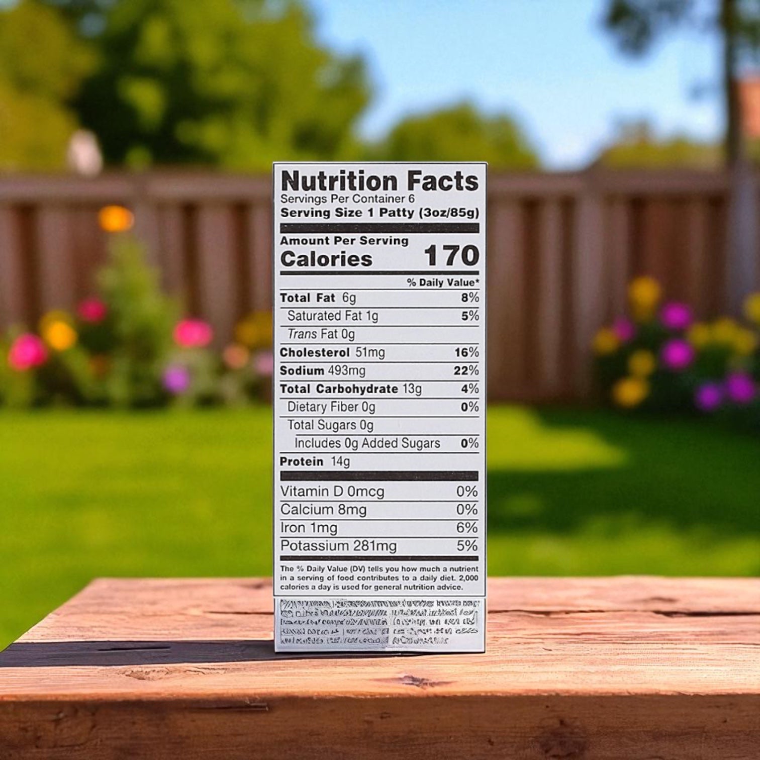 A box of Midamar Halal Chicken Breast Patties (18 OZ) with a nutrition facts label sits outdoors on a wooden surface, grass, flowers, and a wooden fence softly blurred in the background.
