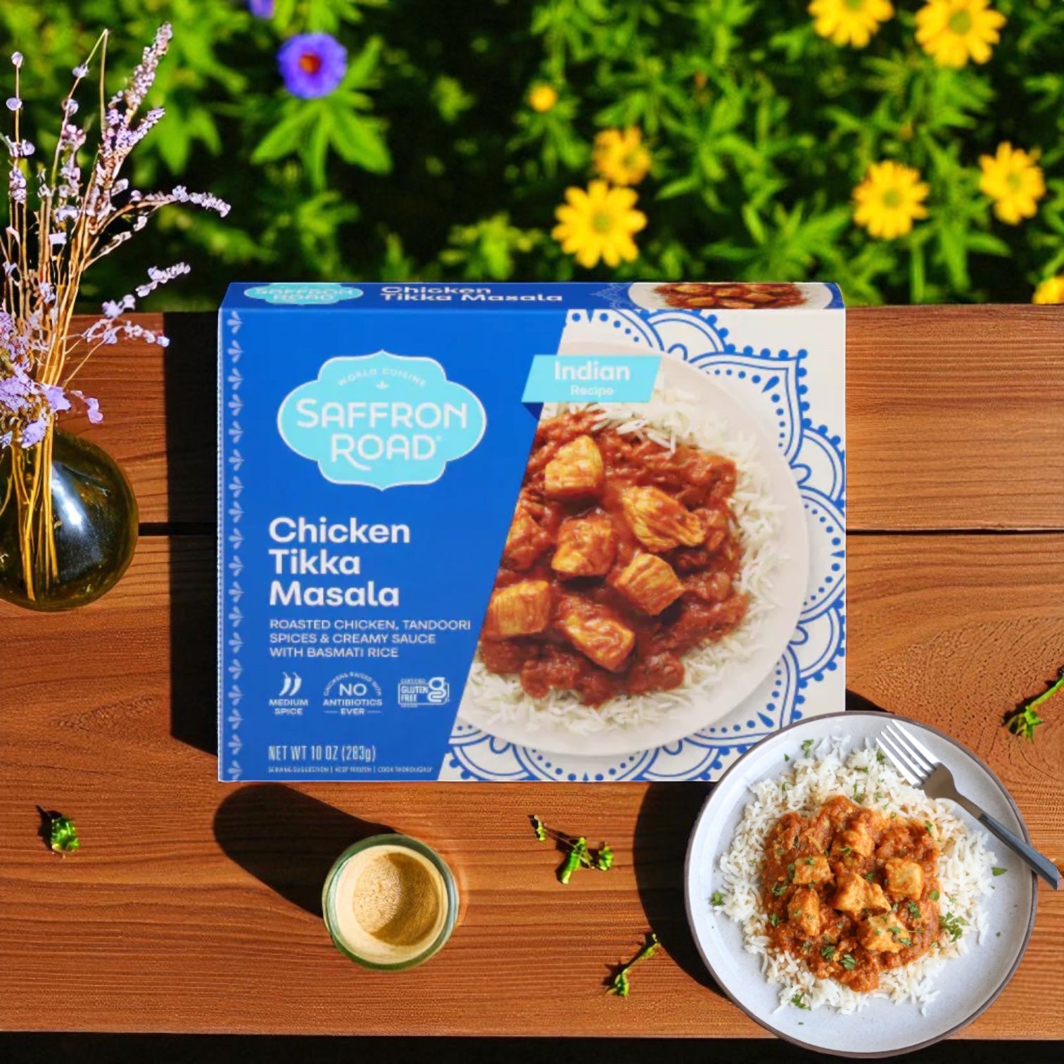 A box of Saffron Road® Chicken Tikka Masala with Basmati Rice Gluten-Free Frozen Dinner (3 Count) sits on a wooden table beside a plated serving, a drink, and vibrant yellow flowers in the background.