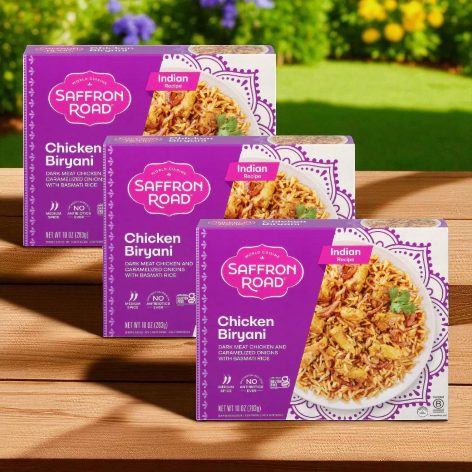 Three boxes of Saffron Road® Chicken Biryani with Basmati Rice Gluten-Free Frozen Dinner (3 Count) are on a wooden surface, highlighting the aromatic rice, with a blurred garden background.