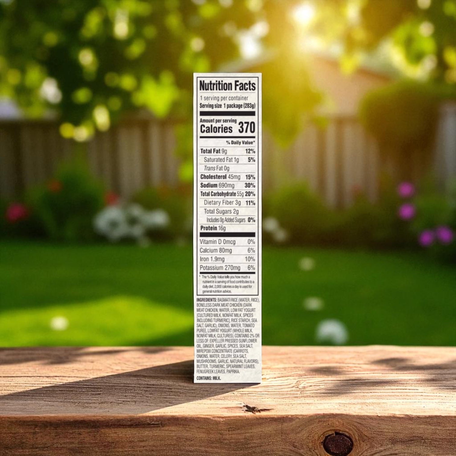 A Saffron Road® Chicken Biryani with Basmati Rice Gluten-Free Frozen Dinner nutrition label is displayed outdoors on a wooden surface, with sunlight highlighting a blurred background of grass, trees, and a fence.