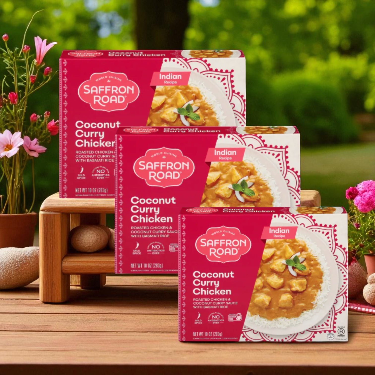 Three boxes of Saffron Road® Coconut Curry Chicken Gluten-Free Frozen Dinner - 3 Count are displayed on a wooden surface outdoors, surrounded by flowers and greenery—a tasty gluten-free meal from Saffron Road.