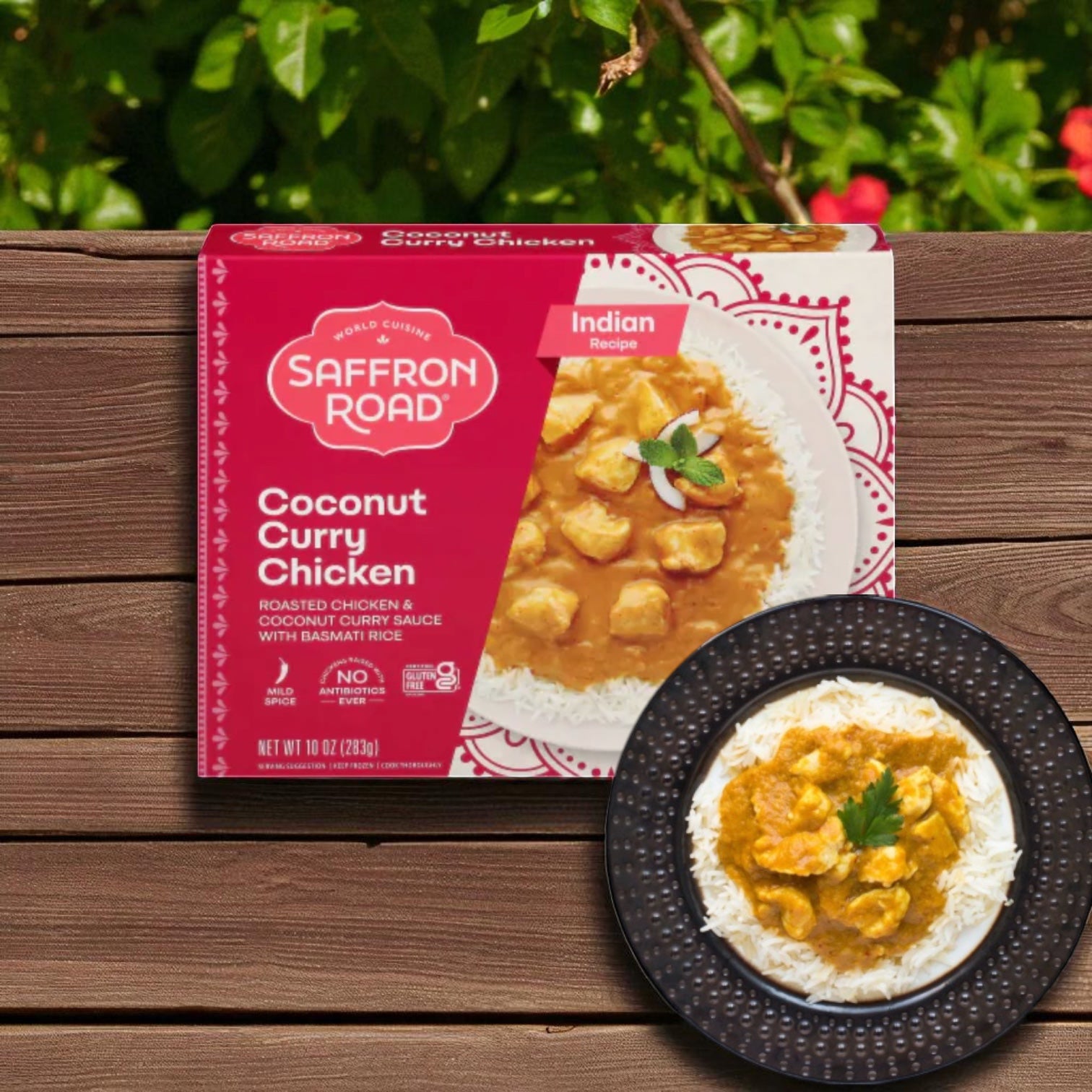 A box of Saffron Road® Coconut Curry Chicken Gluten-Free Frozen Dinner by Saffron Road sits next to a plate of the meal with basmati rice on a wooden surface, with green foliage in the background.