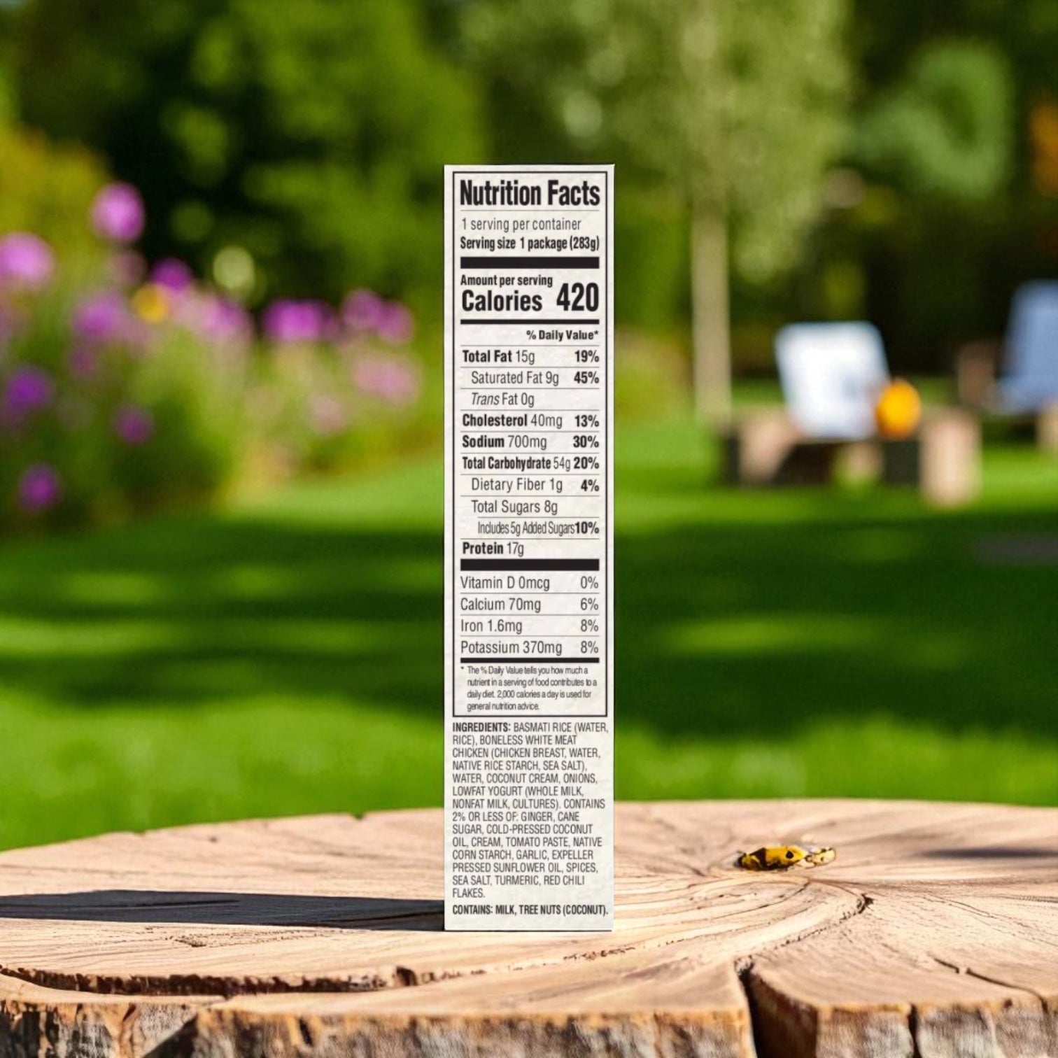 A Saffron Road® Coconut Curry Chicken Gluten-Free Frozen Dinner nutrition label shows 420 calories per serving, standing upright on a wooden surface outdoors with a softly blurred garden in the background.