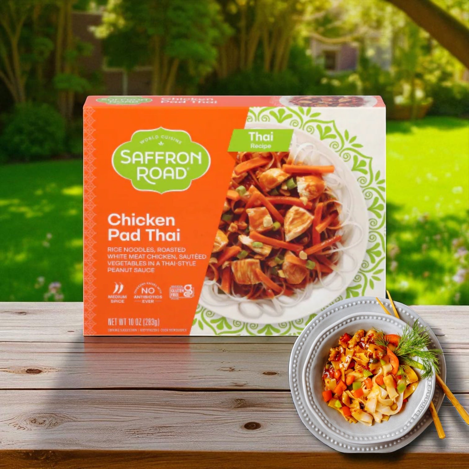 A box of Saffron Road® Chicken Pad Thai with Rice Noodles Gluten-Free Frozen Dinner - 3 Count sits on a wooden table outdoors next to a plated serving, garnished and topped with Thai-style peanut sauce.
