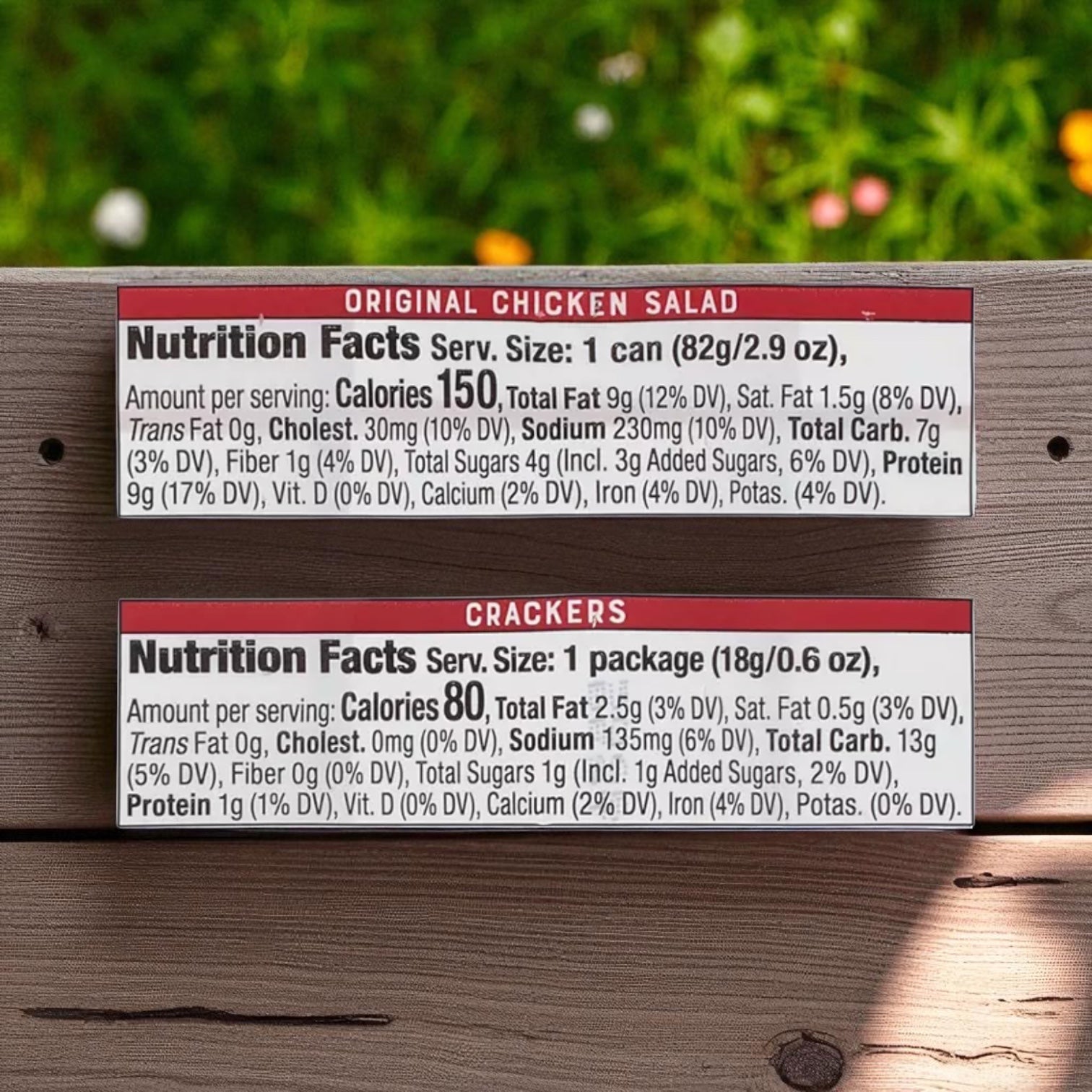 Two nutrition labels for Bumble Bee Snack On The Run Chicken Salad Kits, 3.5 oz., 9 pk., are displayed on a wooden surface with grass and flowers in the background, highlighting this protein-packed snack from Bumble Bee.