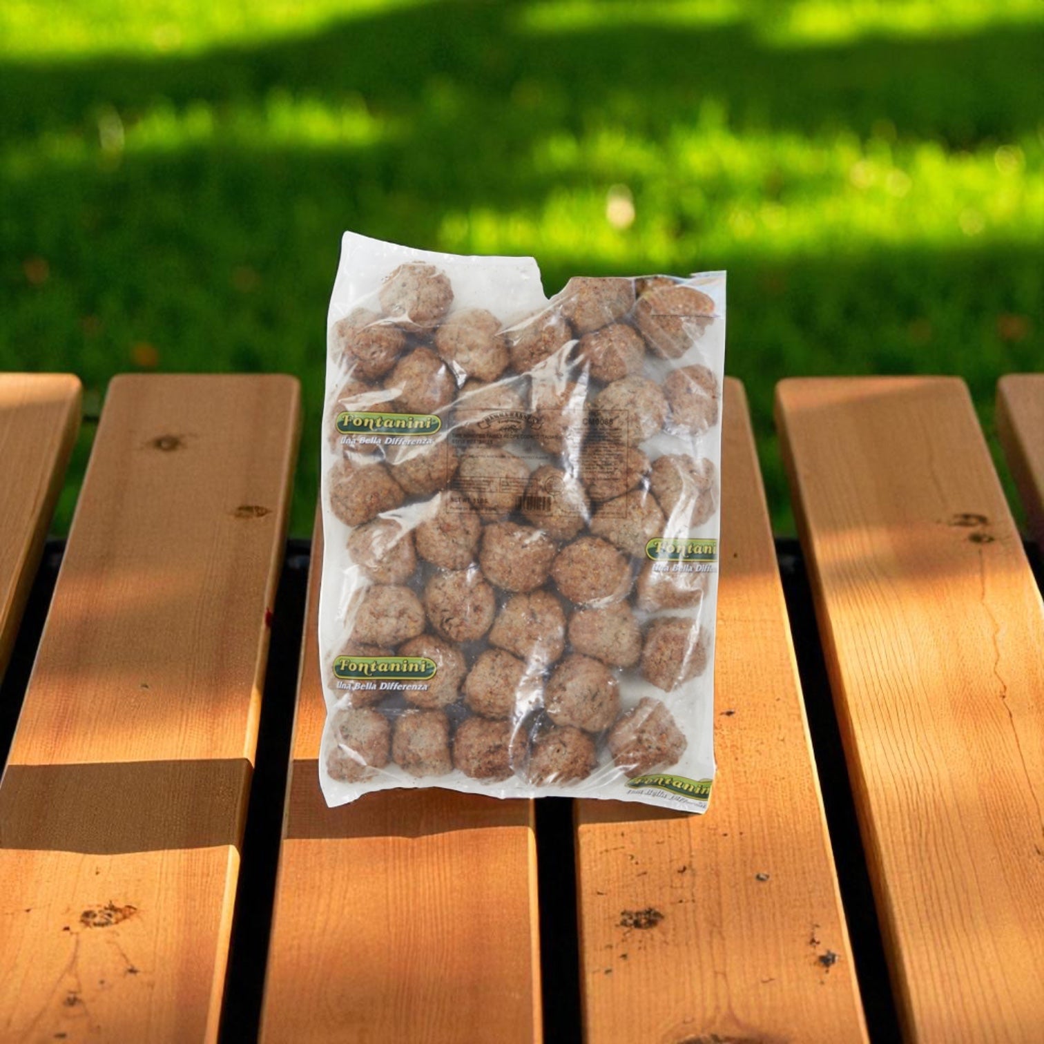 A 5 lb bag of Sysco Fontanini Italian Beef Pork Meatball Fully Cooked 2 Ounce (2 count) sits outdoors on a wooden bench, with green grass in the background.