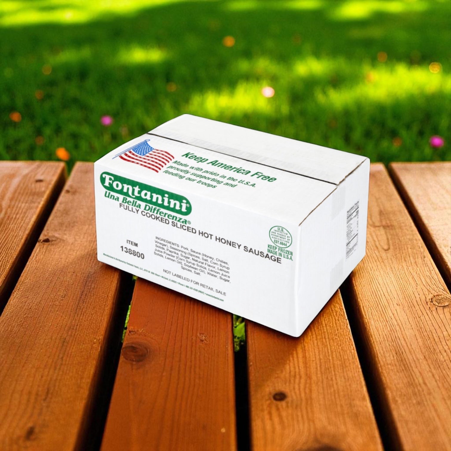 A white box labeled "Sysco Fontanini Italian Honey Cooked Hot Sliced Sausage - 5 lb - 3 Count" rests on a wooden table outdoors, with grass and sunlight in the background.