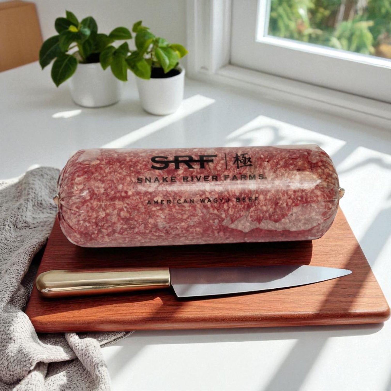 A 5 lb log of Snake River Farms American Style Wagyu Beef Ground (75%, 4 Count) rests on a wooden cutting board beside a knife, with a houseplant and window visible in the background.