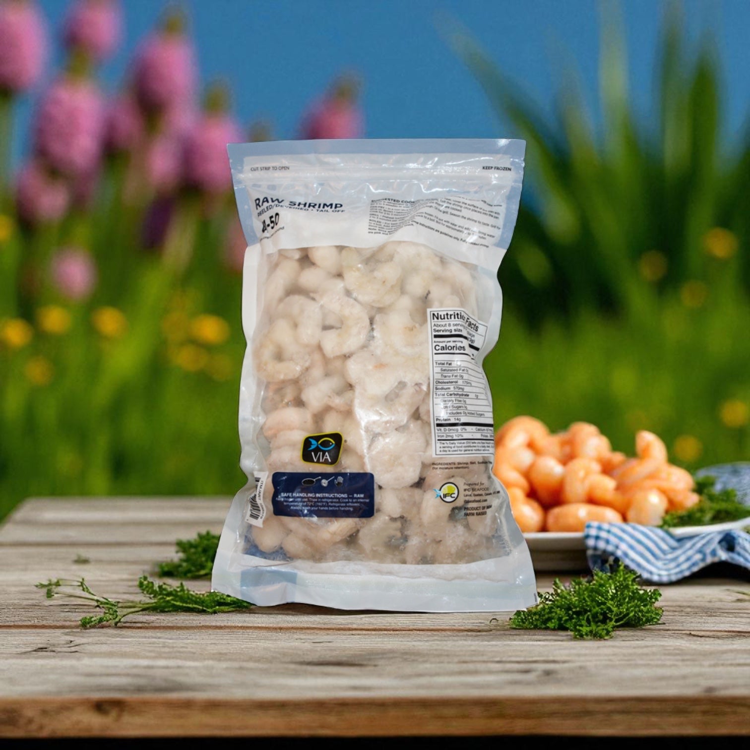 A sealed bag of Via Farms Farm-Raised Raw Shrimp (Tail-Off, Peeled, Deveined, 41-50 ct, 2 lbs) sits on a wooden table outdoors beside a plate of shrimp and fresh herbs, with blurred greenery and flowers in the background.