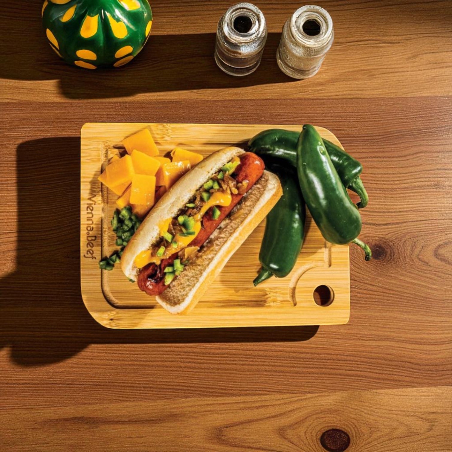 A wooden cutting board displays a Vienna Beef Cheddar Frank from the 12 oz 12 pack, topped with onions, relish, cheddar cheese slices, diced green peppers, and whole jalapeños on a table with salt and pepper shakers.