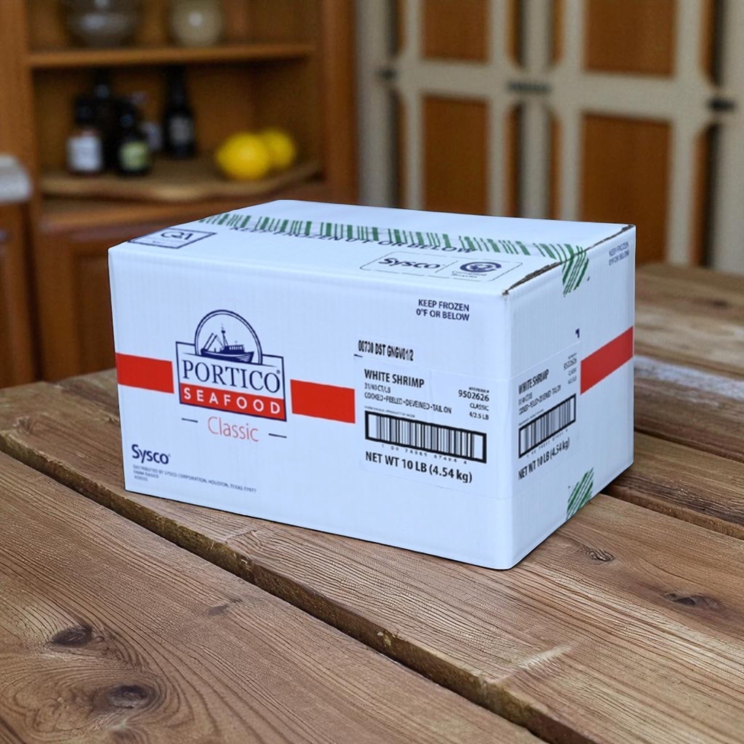 A white Sysco box labeled "Shrimp White Peeled And Deveined Tail-on Cooked 2.5 lb, 4 Count" sits on a wooden kitchen table, featuring convenient peeled and deveined tail-on shrimp.